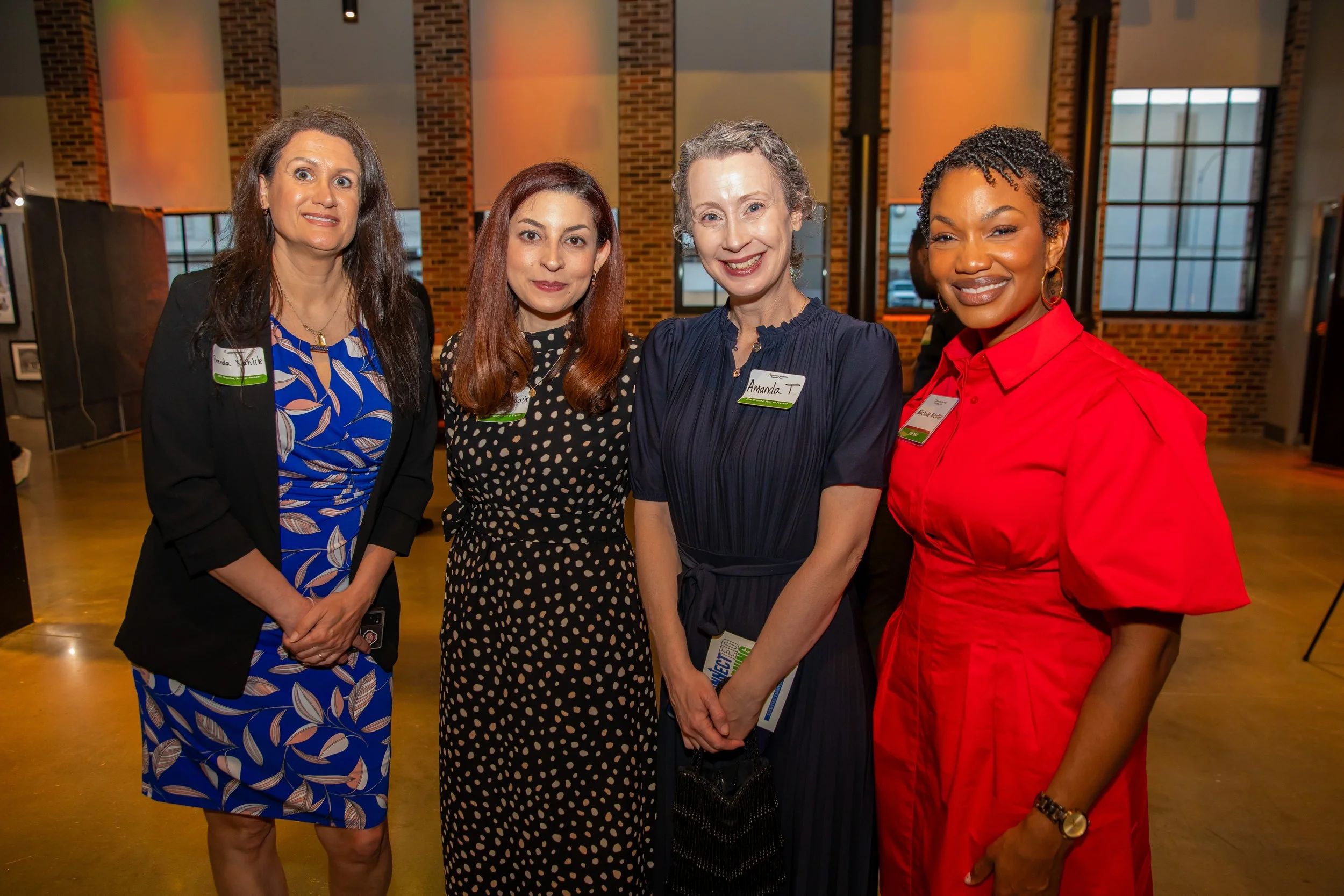 Four women standing together indoors, smiling for a photo at a professional event. They are wearing name tags and dressed in business or semi-formal attire.