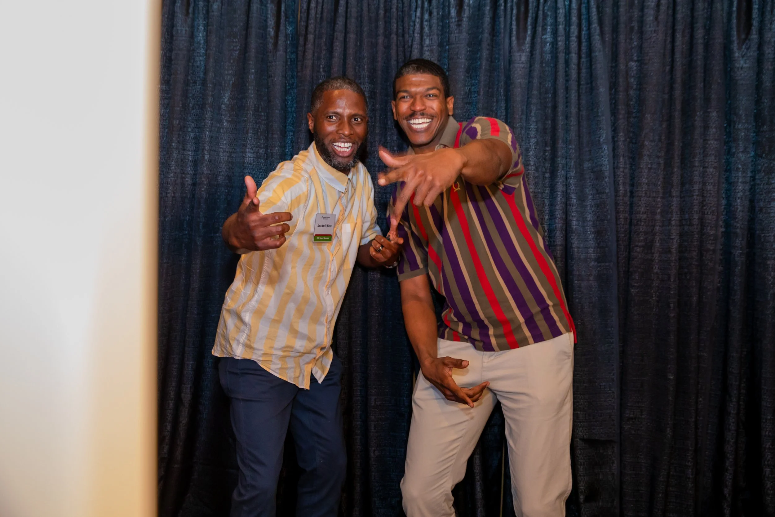 Two men smiling and posing joyfully in front of a dark curtain, with one man pointing towards the camera and the other holding his hand in a fist.