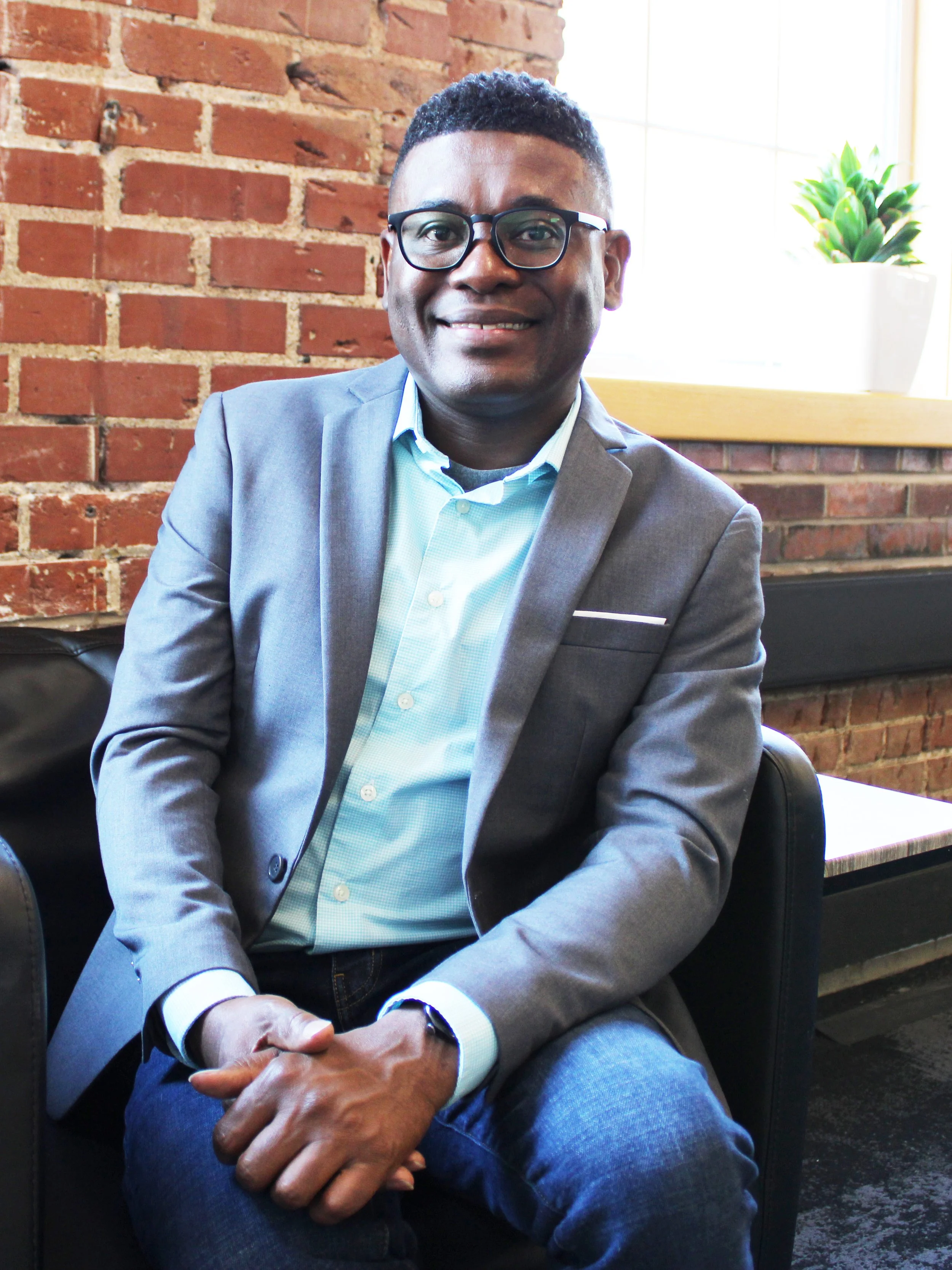 A man wearing a gray blazer, light blue shirt, and glasses sitting in front of a brick wall with a window and a potted plant in the background.