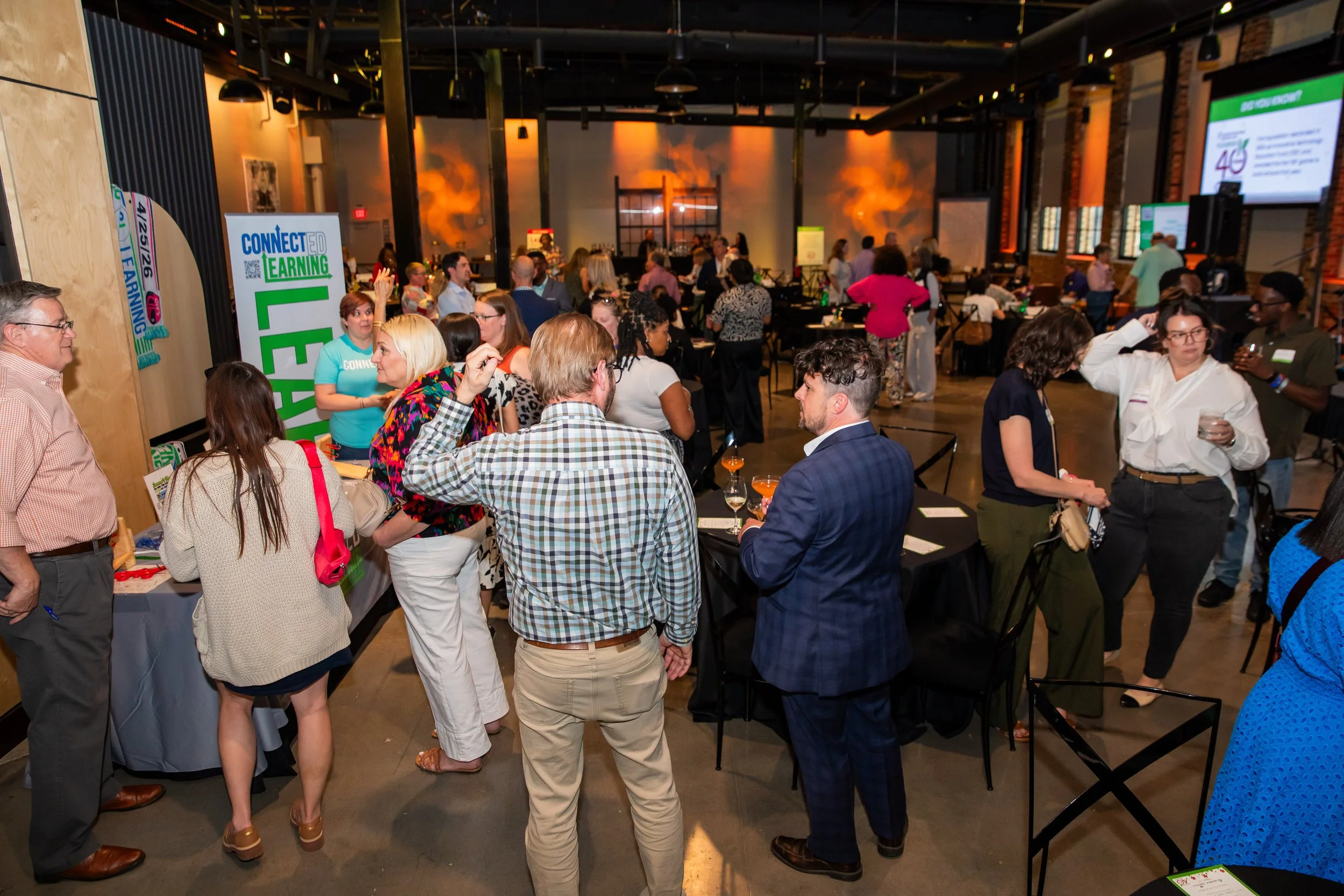 People gathered at a conference or networking event in a large, industrial-style venue with high ceilings, exposed brick, and multiple screens displaying information.
