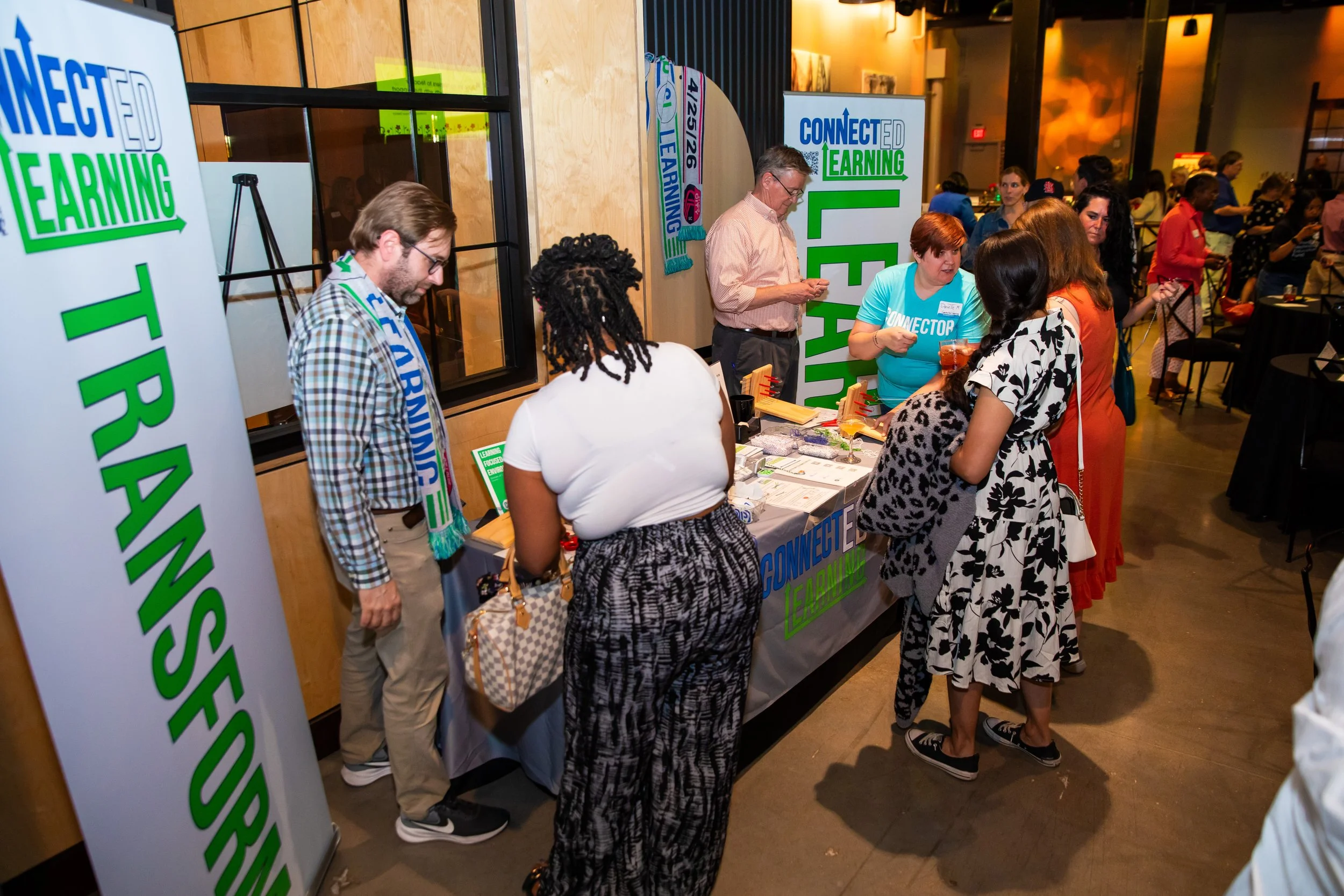 People attending a conference registration or check-in booth for Connected Learning, with banners and tables displaying materials, and individuals engaging with staff.