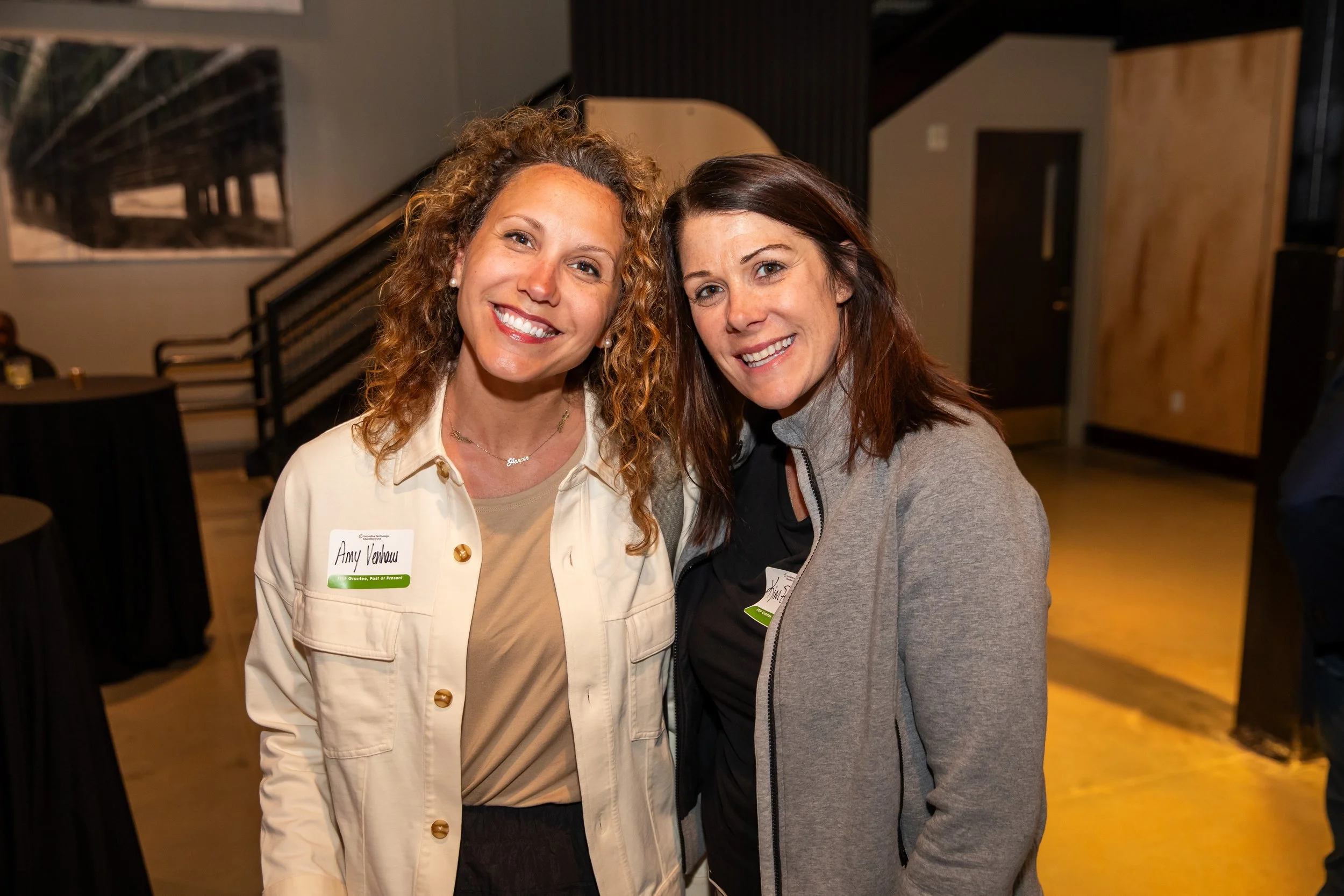 Two women smiling at the camera indoors. The woman on the left has curly red hair, wears a beige jacket, and has a name tag. The woman on the right has straight brown hair, wears a gray zip-up jacket, and also has a name tag.