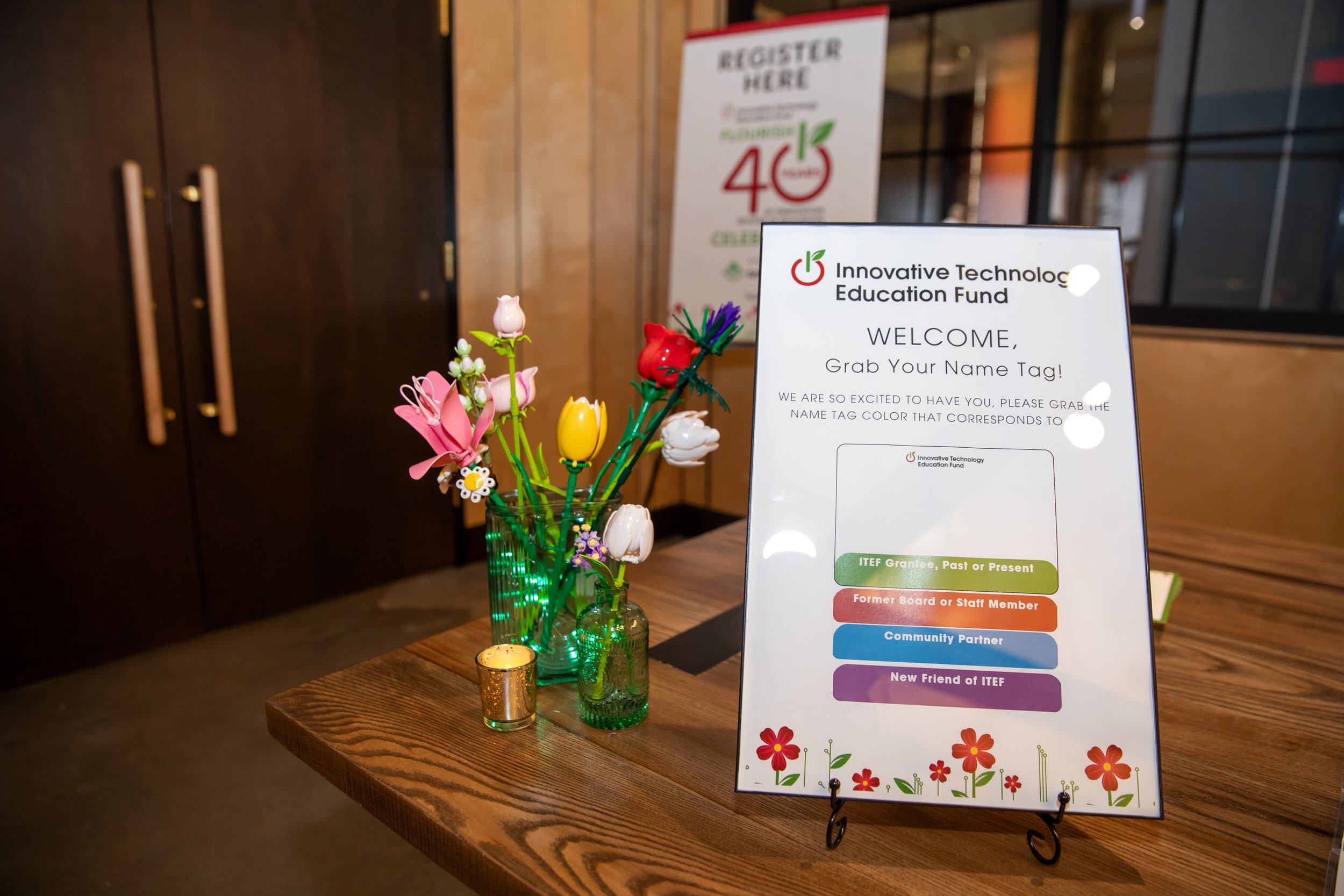 Decorative table with pink and white flower arrangements in green glass vases, a small gold votive candle holder, and a sign for the Innovative Technology Education Fund registration with colorful sections and a floral border.