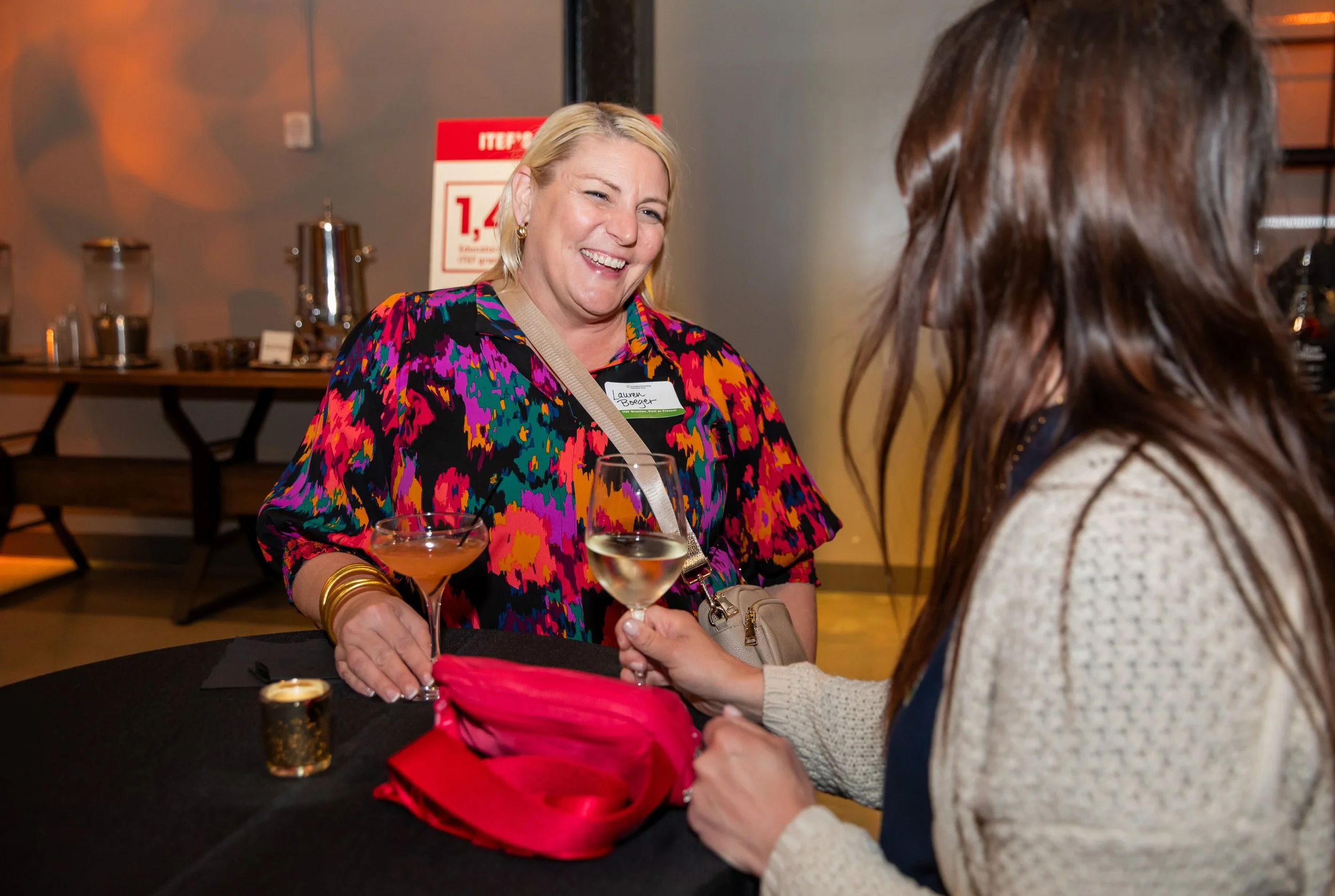 Two women at a social event, one with blonde hair wearing a colorful patterned blouse and gold jewelry, smiling and holding a cocktail, and the other with brown hair, wearing a beige sweater, holding a drink. In the background, buffet table with serv