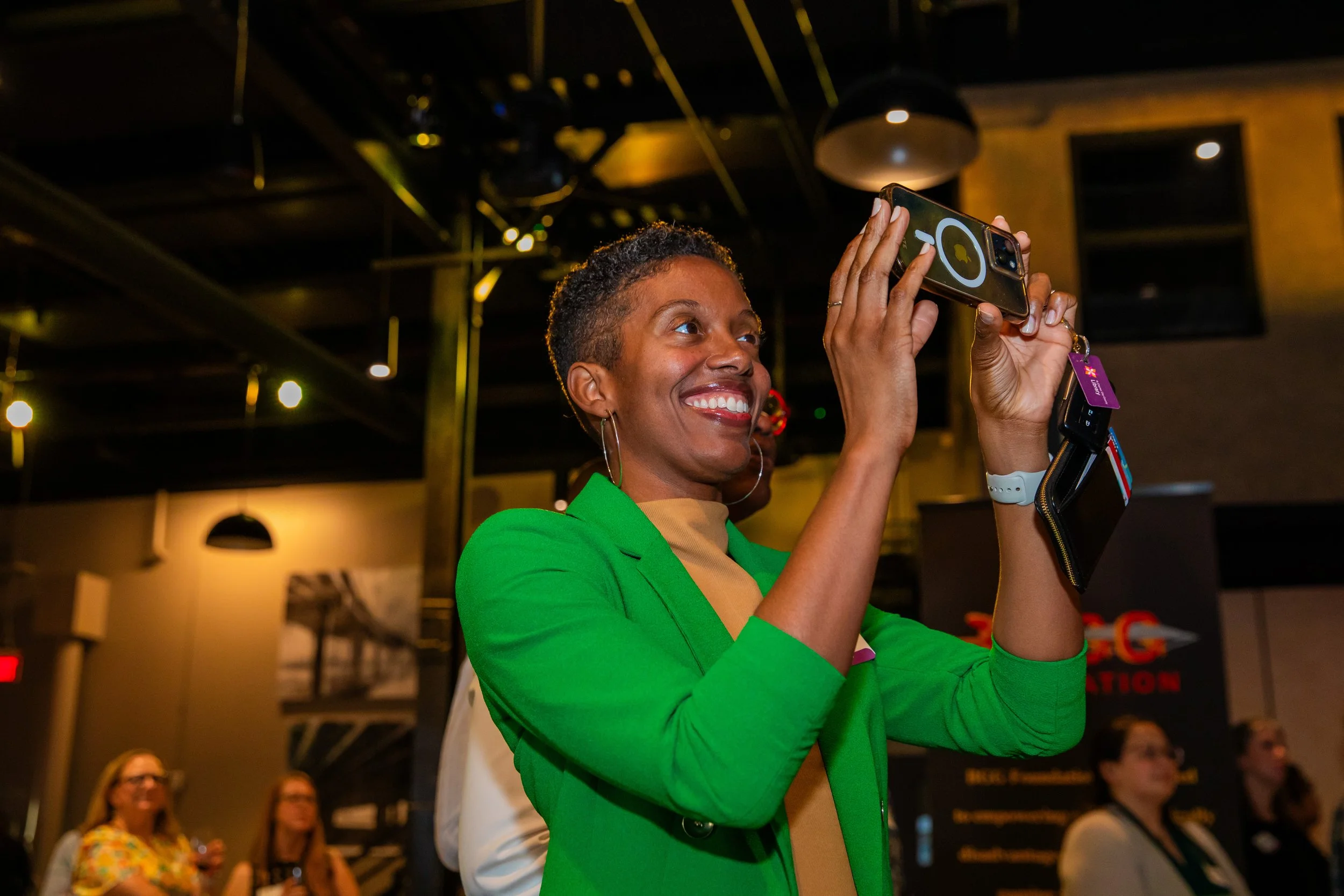 A woman with short curly hair and a bright smile is taking a photo with her smartphone in a lively indoor setting. She is wearing a vibrant green blazer. Other people are seated and watching in the background.