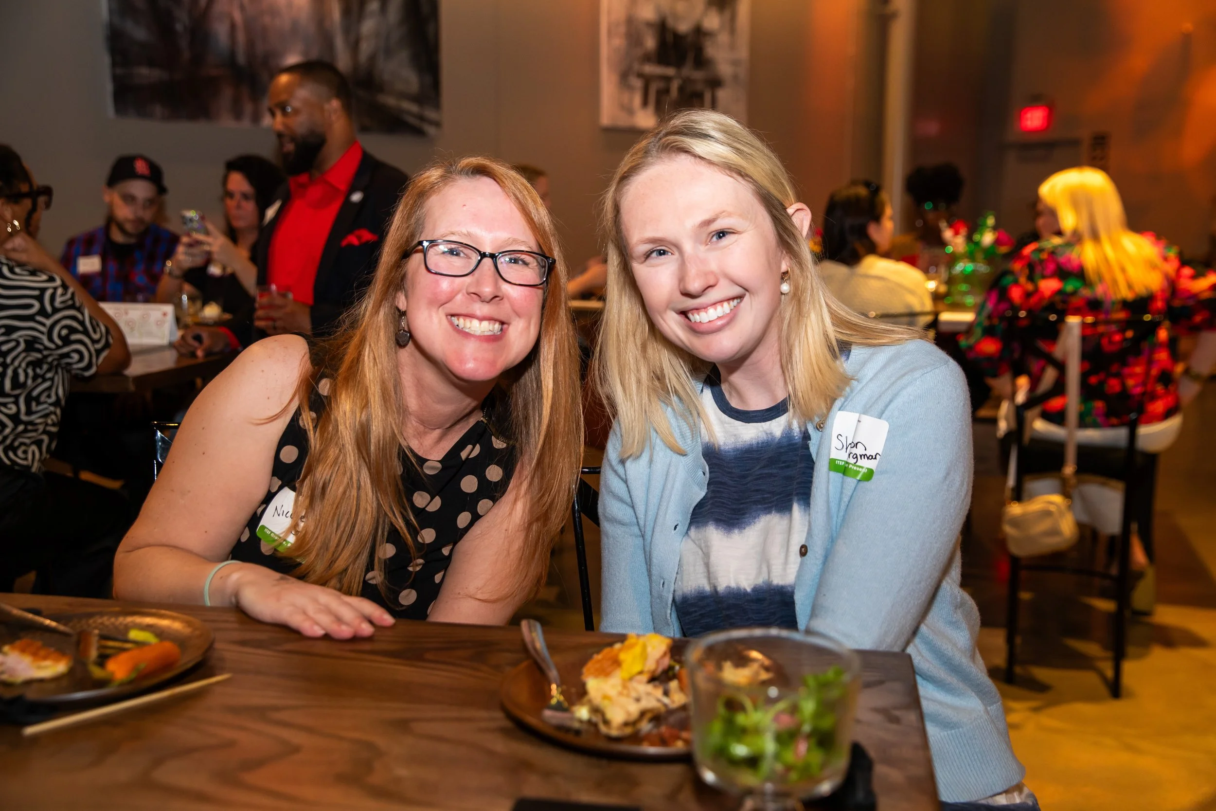 Two women smiling at a social event, sitting at a table with food and drinks, with people in the background in a dimly lit room.