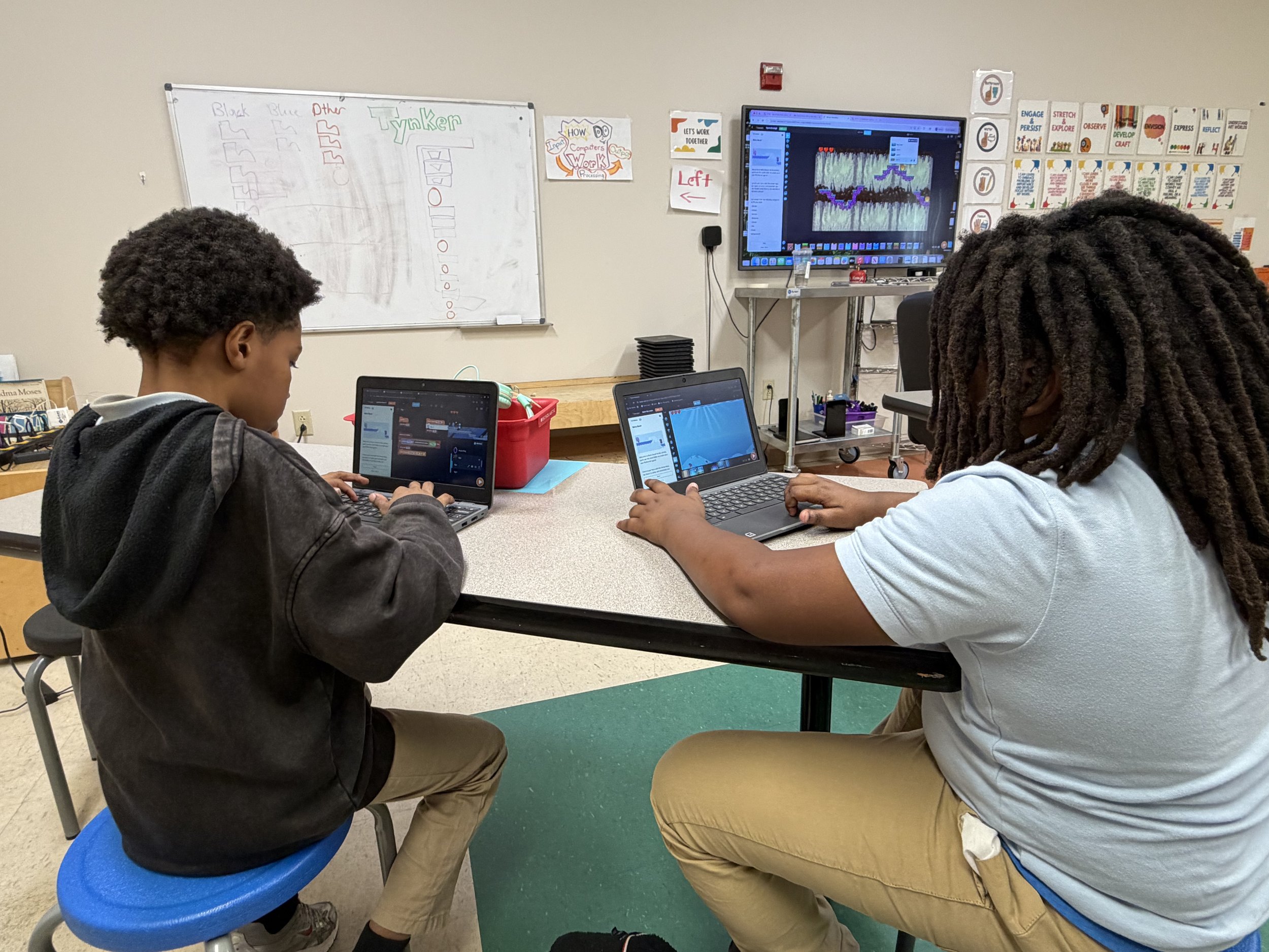 Two students sitting at a table working on laptops in a classroom. Behind them, a whiteboard displays writing and diagrams, and a large monitor shows a digital game or animation with a colorful landscape.