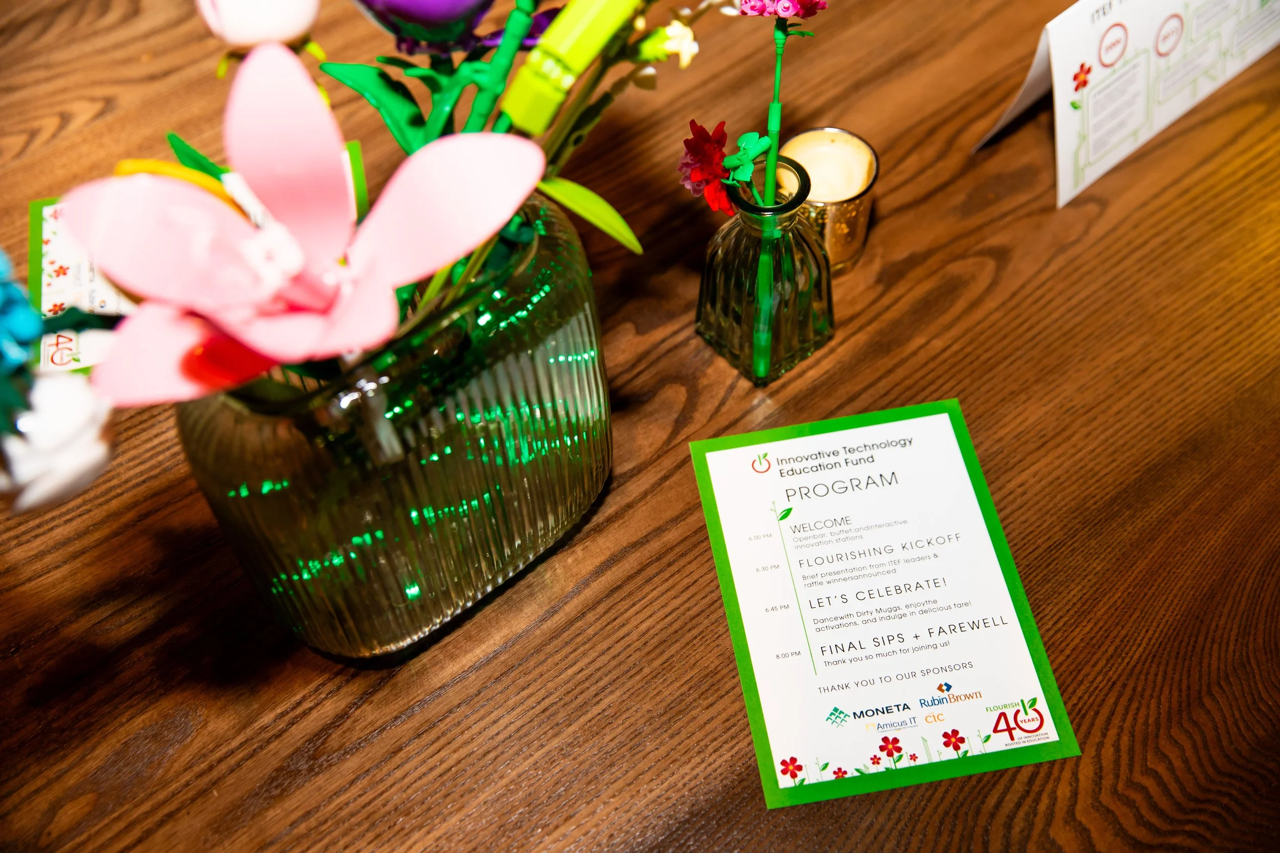 A wooden table with a green glass vase holding pink paper and plastic flowers, next to a small glass vase with green plastic flowers. On the table, there is a program sheet with a schedule of events, including welcome, flourishing kickoff, celebratio