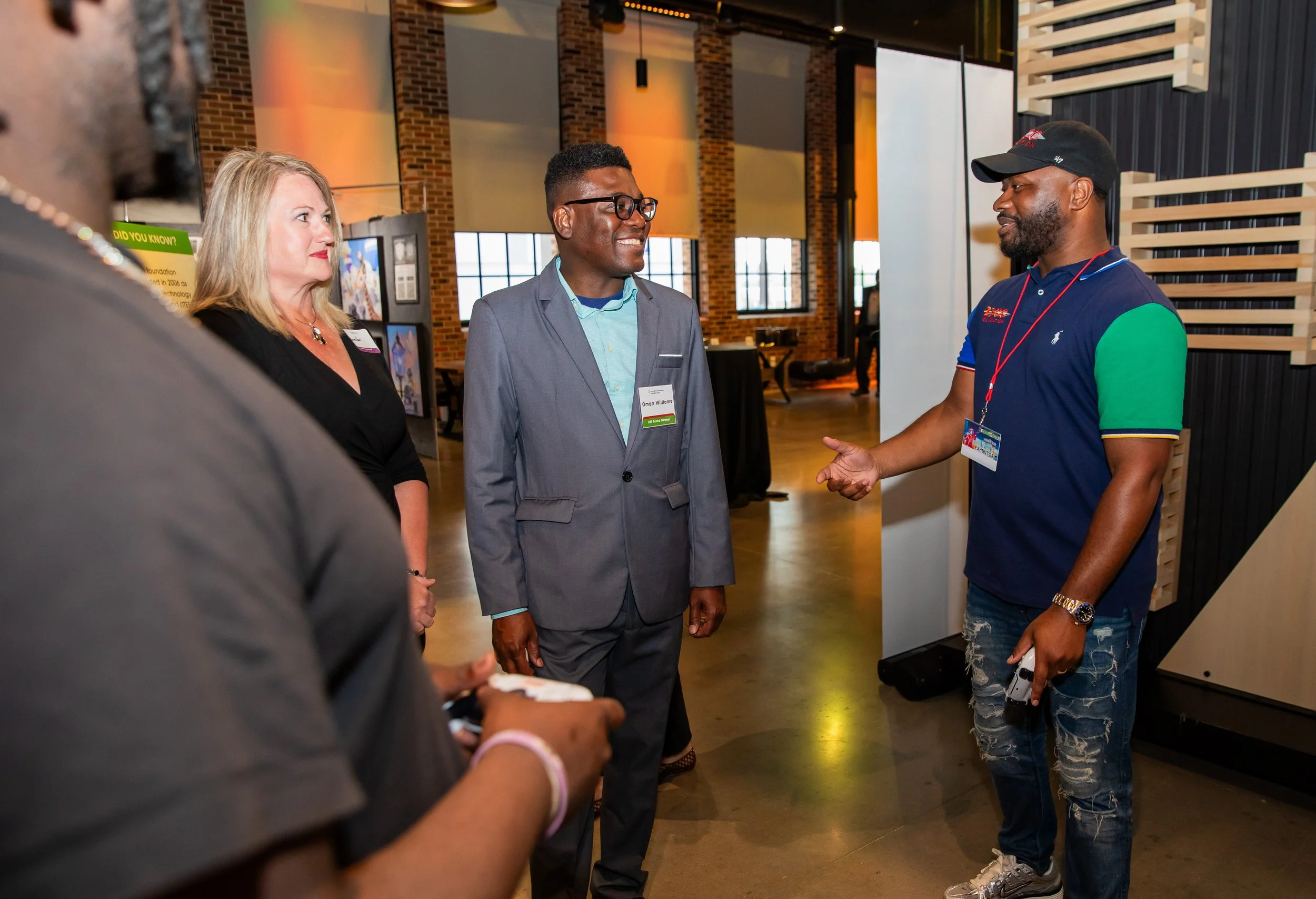 Three people are engaged in conversation at an indoor event. One man, in a gray blazer and glasses, faces a young man in a colorful polo shirt and a baseball cap. A woman with blonde hair and dressed in black is also present, partially visible on the