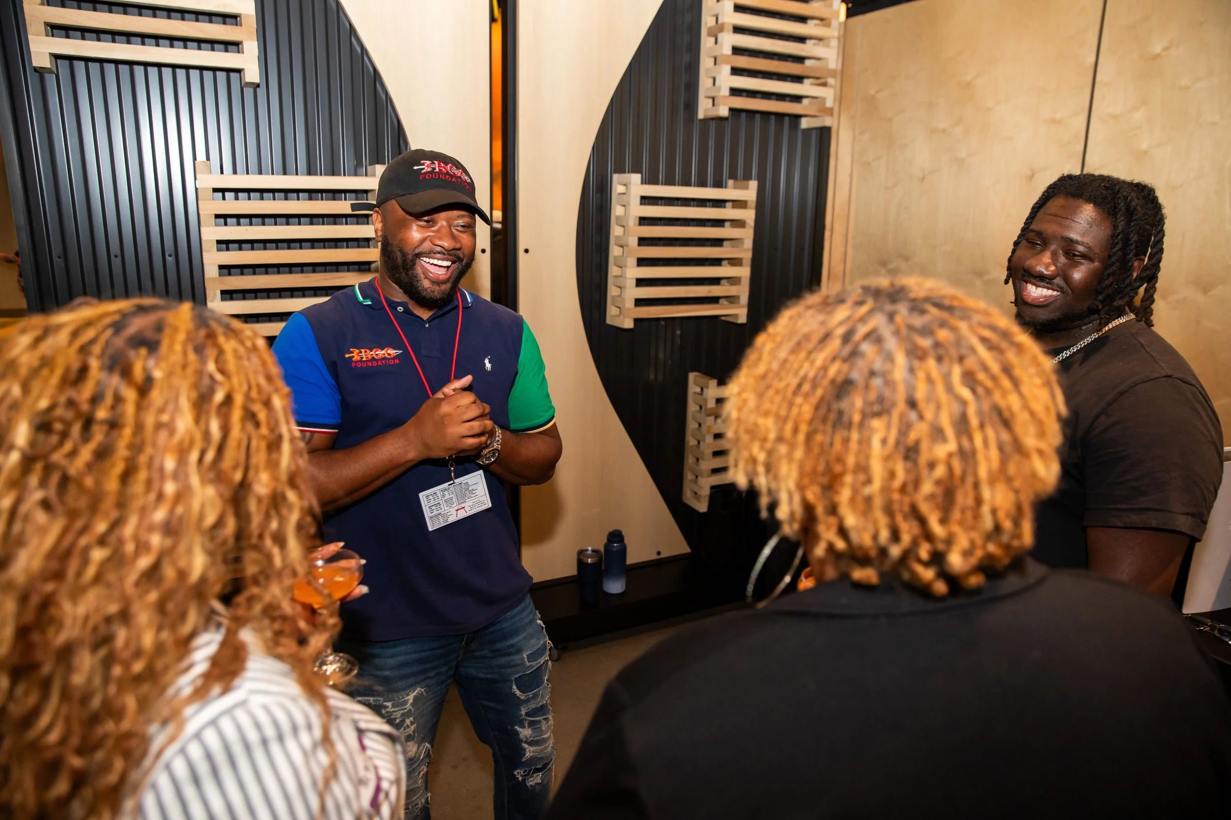 A group of people gathered around two men smiling and talking indoors, with wood and black panel decor behind them. One man is wearing a cap and a polo shirt, the other has dreadlocks and a necklace.