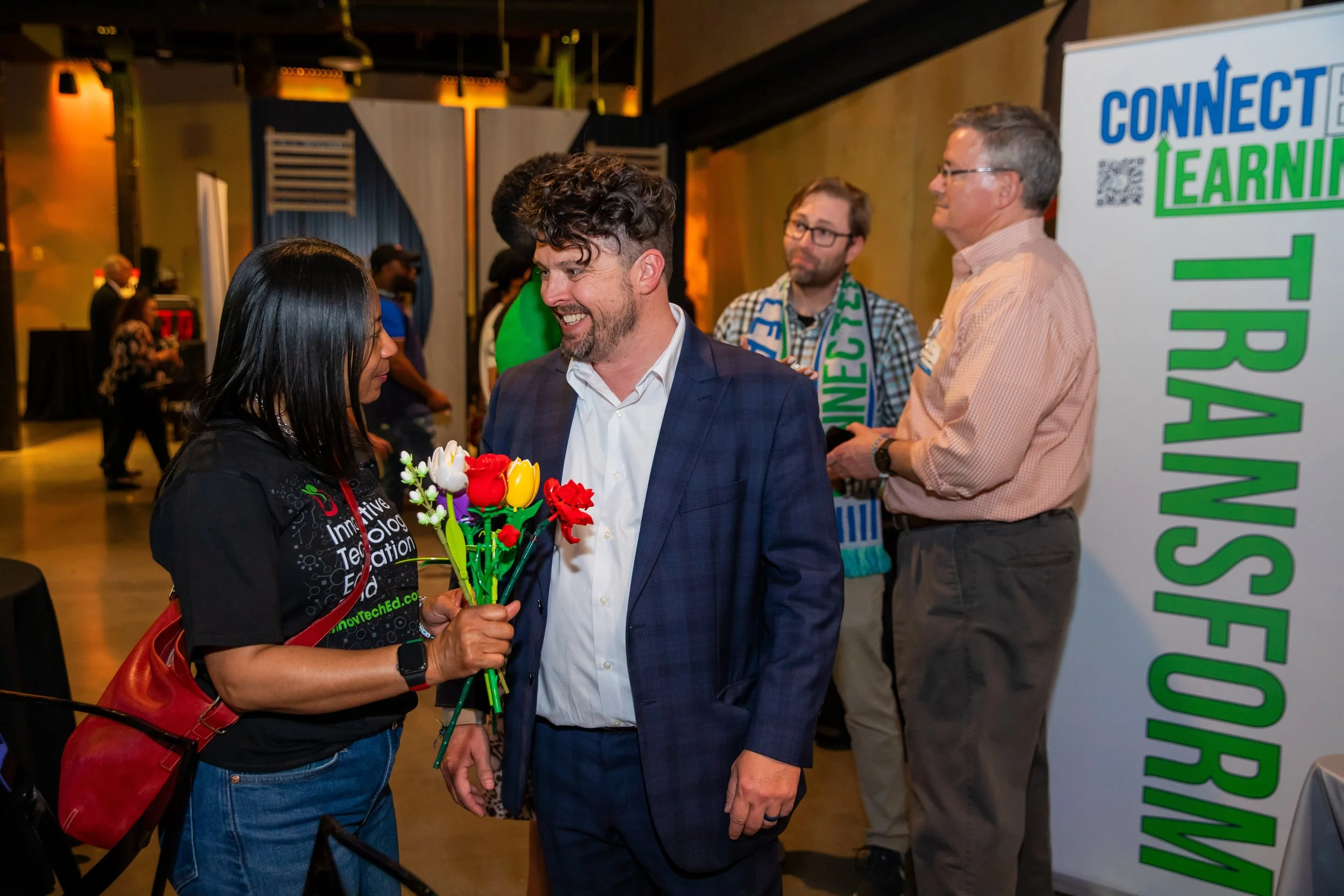 A woman giving flowers to a man in a suit at an event, with a banner in the background that says 'Connect, EARN, TRUST'.