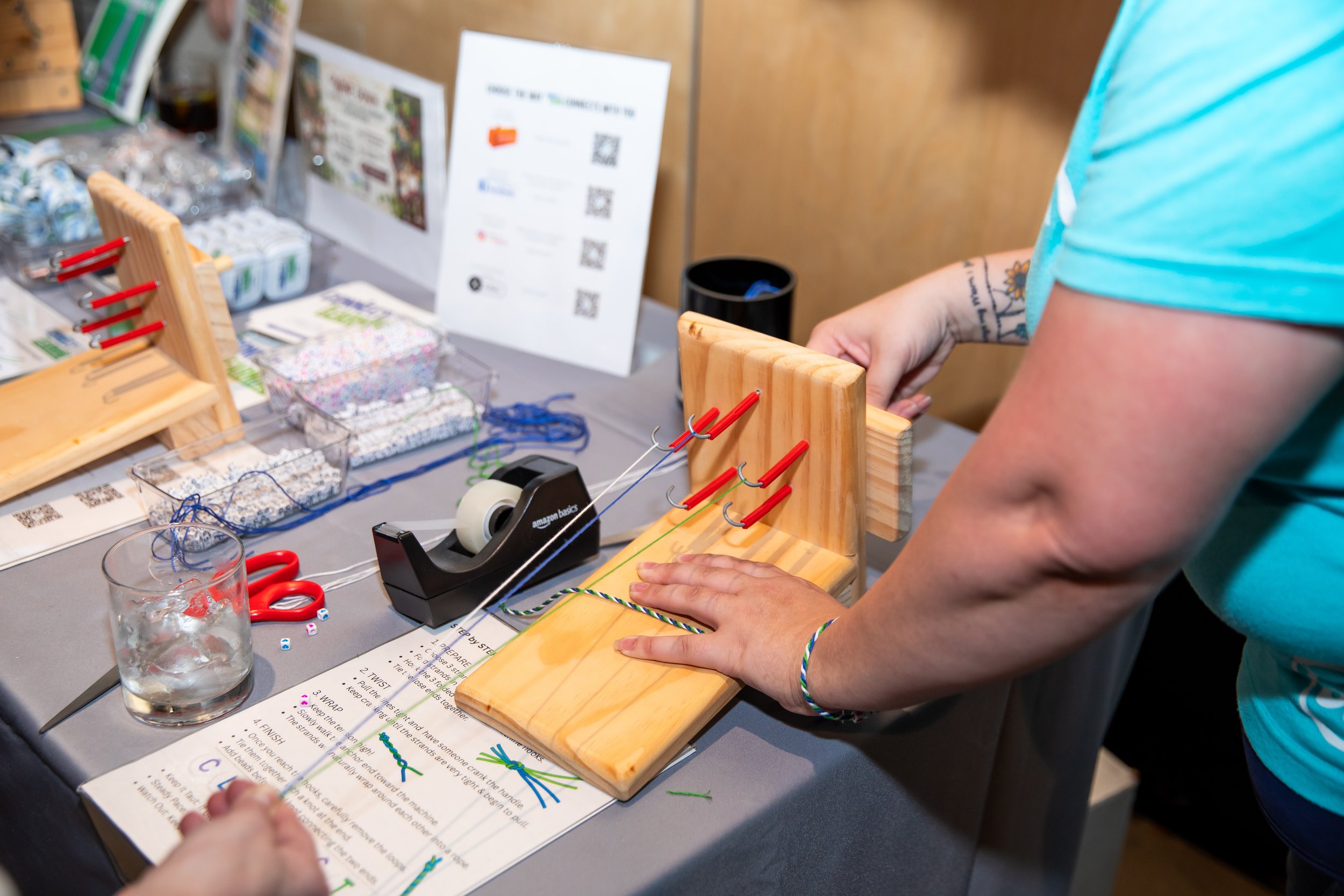 Person demonstrating how to tie dye using a wooden stand with colorful strings, with various craft supplies and instruction sheet on the table.