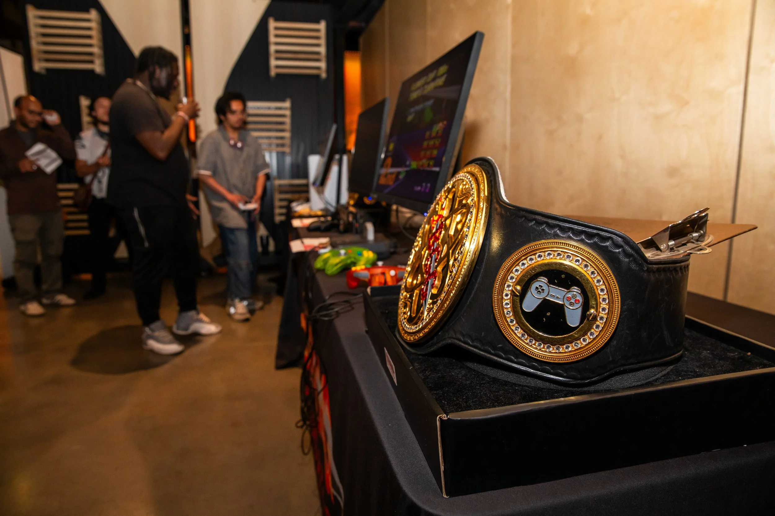 A championship belt with a gaming controller icon in the center, decorated with rhinestones, placed on a table at a gaming event. In the background, a group of people are gathered around a computer screen.