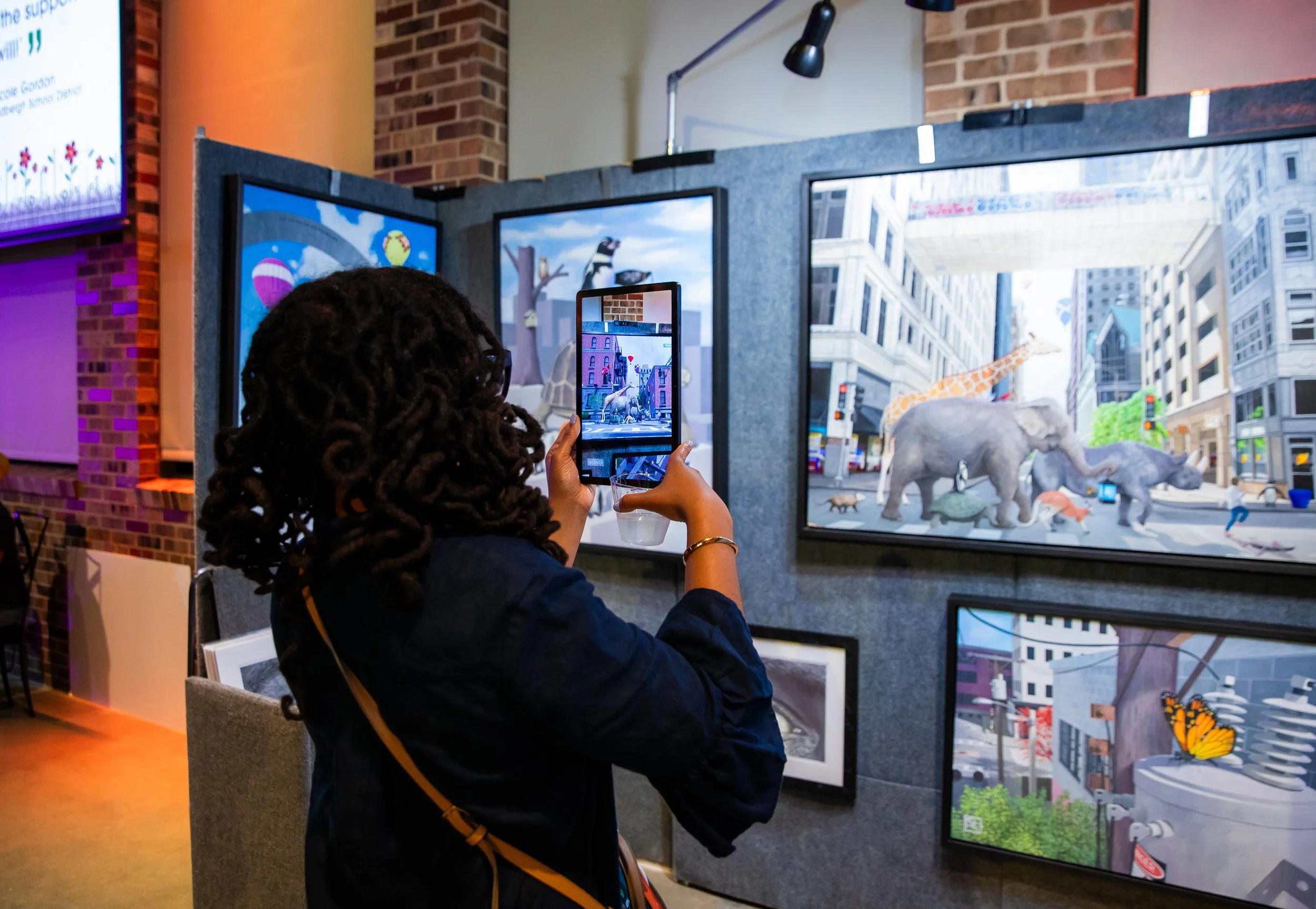 A woman with curly hair taking a photo of a digital display with her phone. The display shows animated animals, including elephants, giraffes, and other wildlife, in an urban setting inside a building.