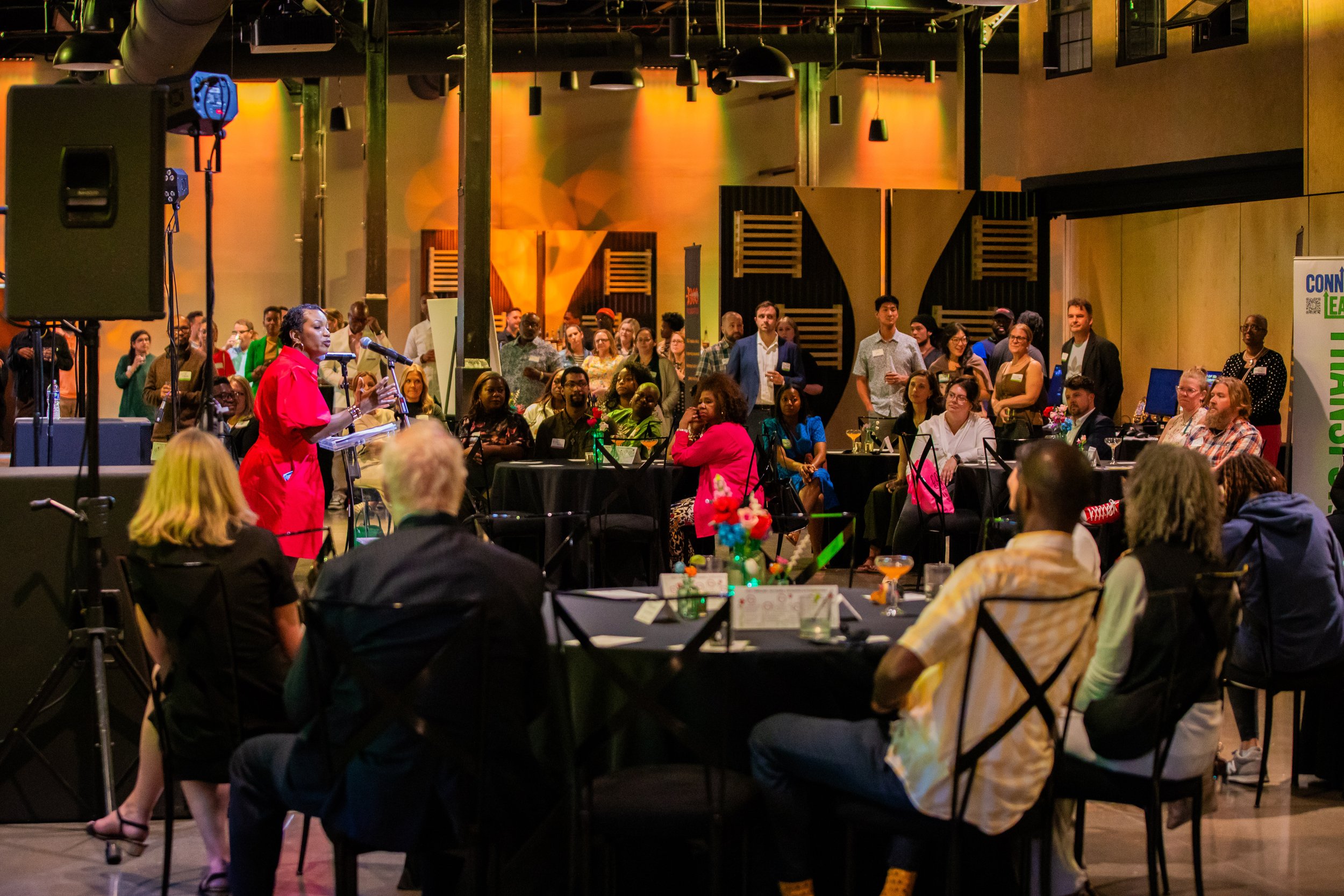 A woman in a red outfit speaking into a microphone at a conference or event, with an audience seated at round tables in a dimly lit decorated venue.