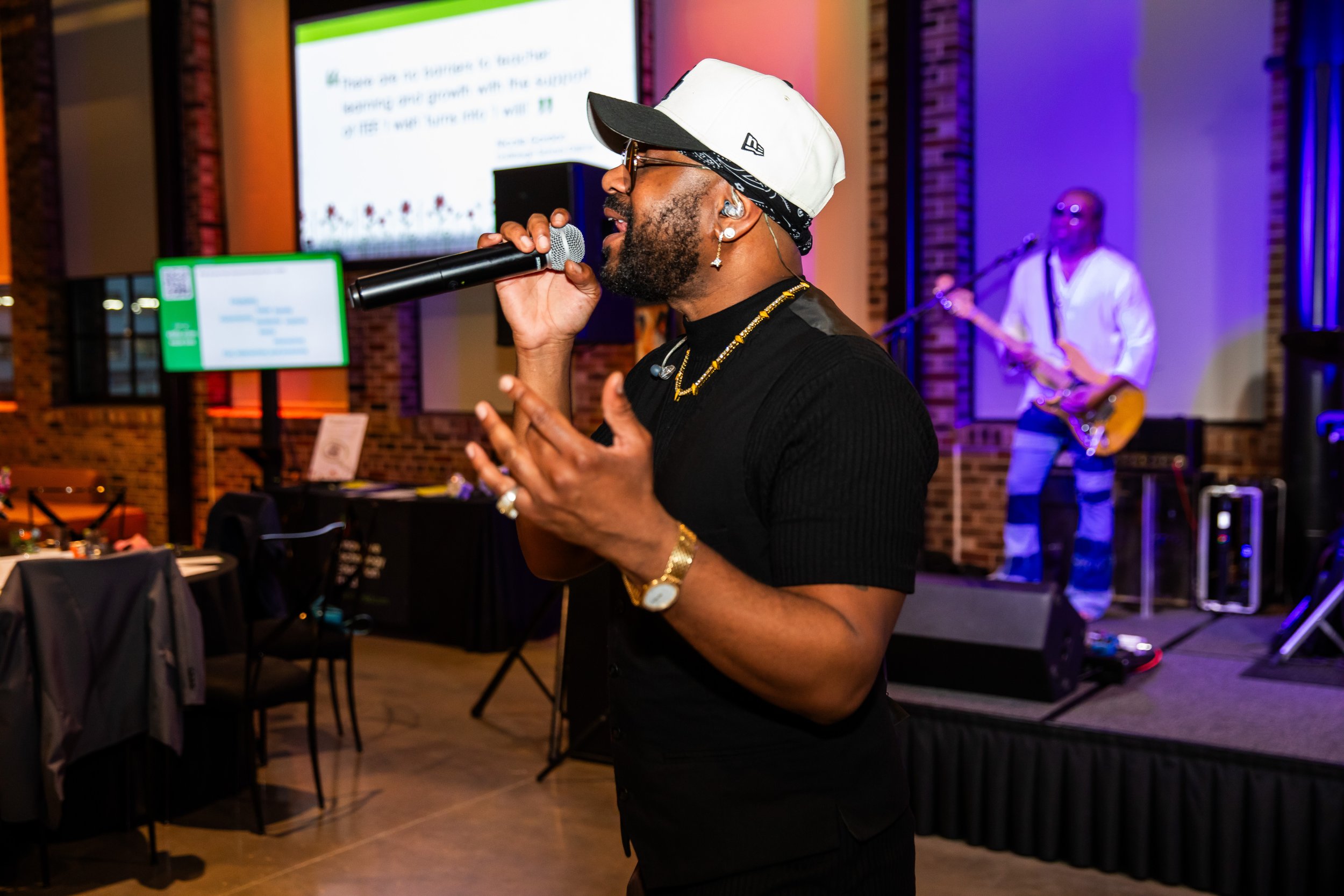 A man singing into a microphone during a performance at an indoor event with a live band in the background. The man is wearing a white cap, black shirt, glasses, and jewelry. A large screen and tables are visible in the background.