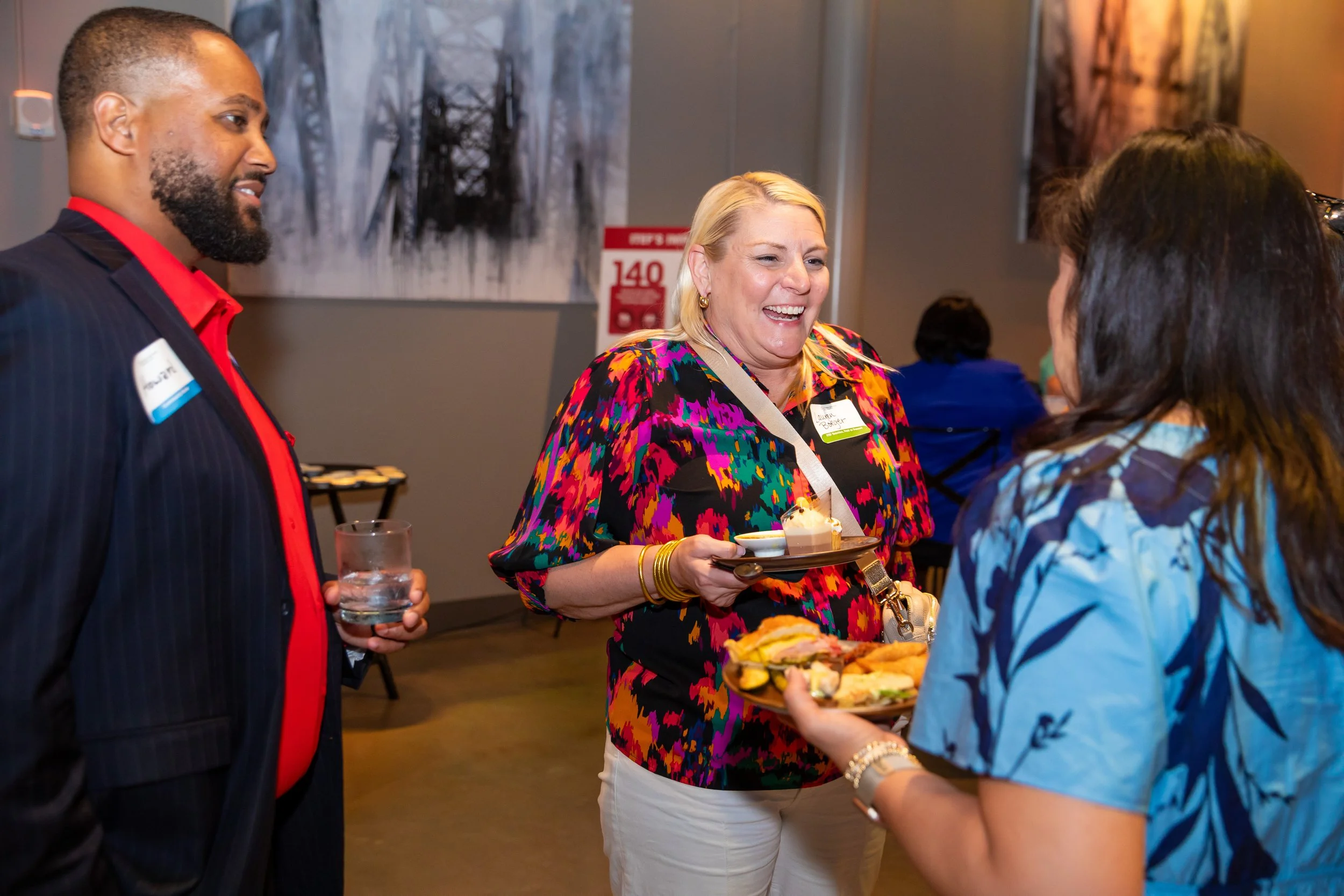 Three people are at a social event talking and sharing food. The woman in the colorful blouse is smiling and holding a tray of appetizers, while a man in a suit and red shirt and a woman in a blue patterned shirt are engaged in conversation with her.