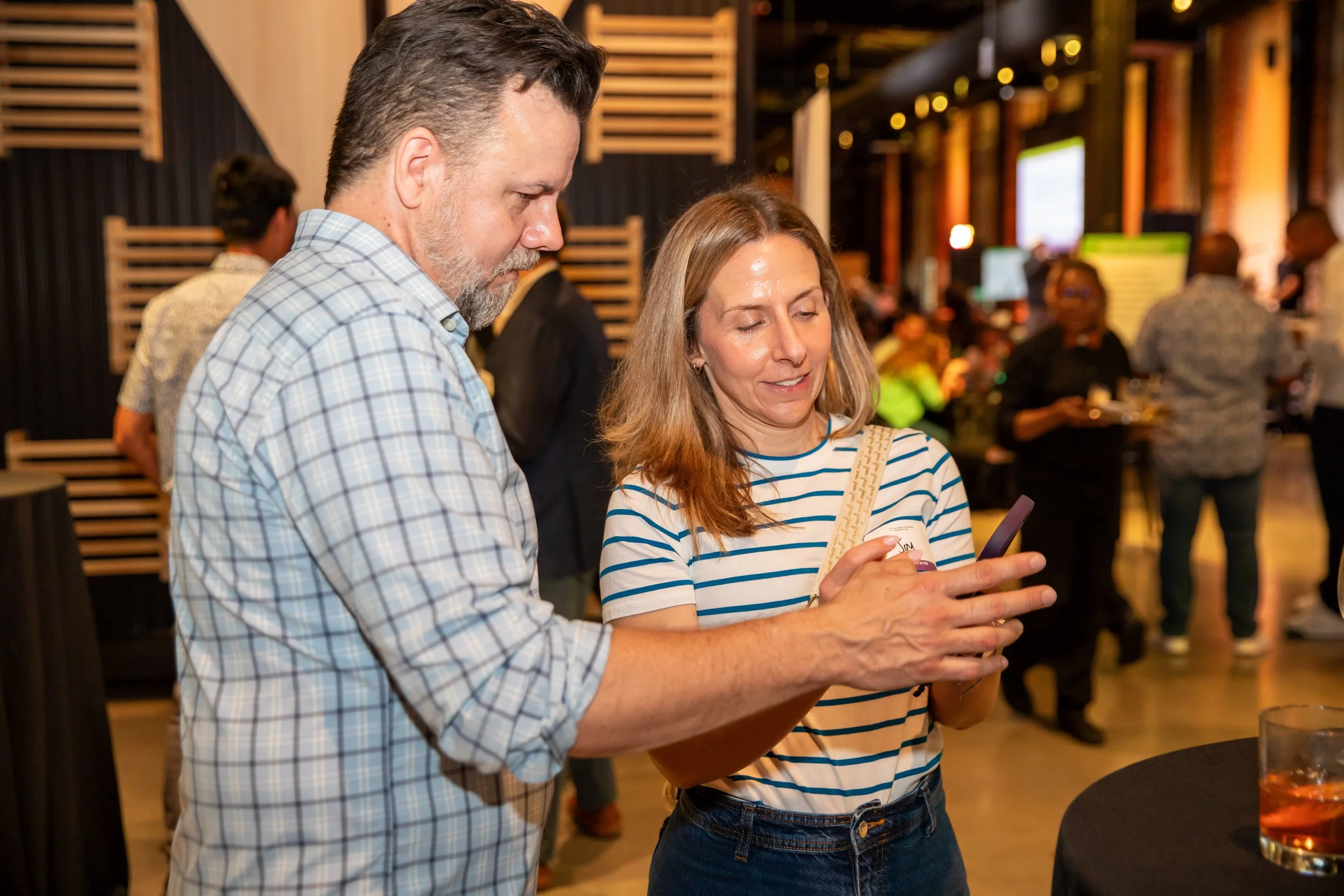 A man with gray hair and a woman with long blonde hair are looking at a smartphone together at a social event. The woman is smiling and holding the phone, wearing a striped shirt, and the man is wearing a plaid shirt. The background shows other peopl