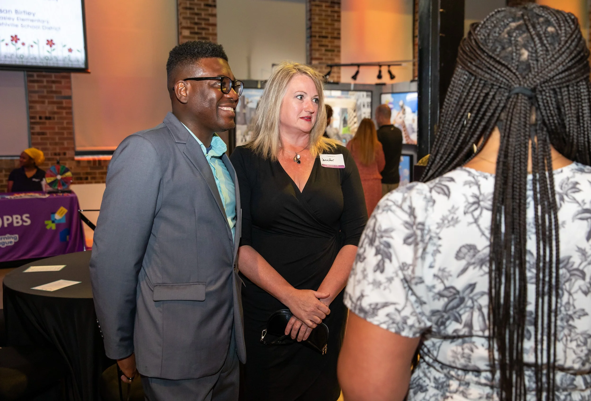 Three people are engaged in conversation at an indoor event. The man on the left is smiling and wearing glasses and a light gray suit. The woman in the middle has blonde hair, is wearing a black dress, and has a name tag that says 'Jennifer.' The per