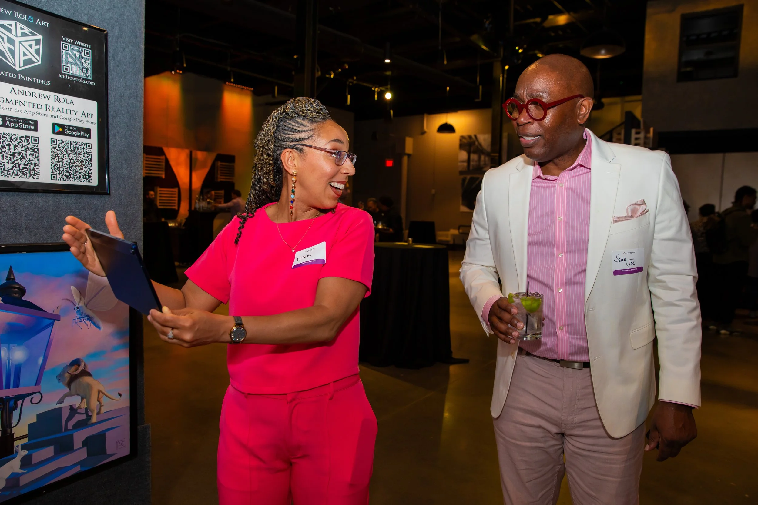Two people, a woman with glasses and curly hair in a pink outfit and a man with glasses in a white blazer and pink shirt, are smiling and talking at an indoor event. The woman is holding a tablet, and the man is holding a drink with lime.