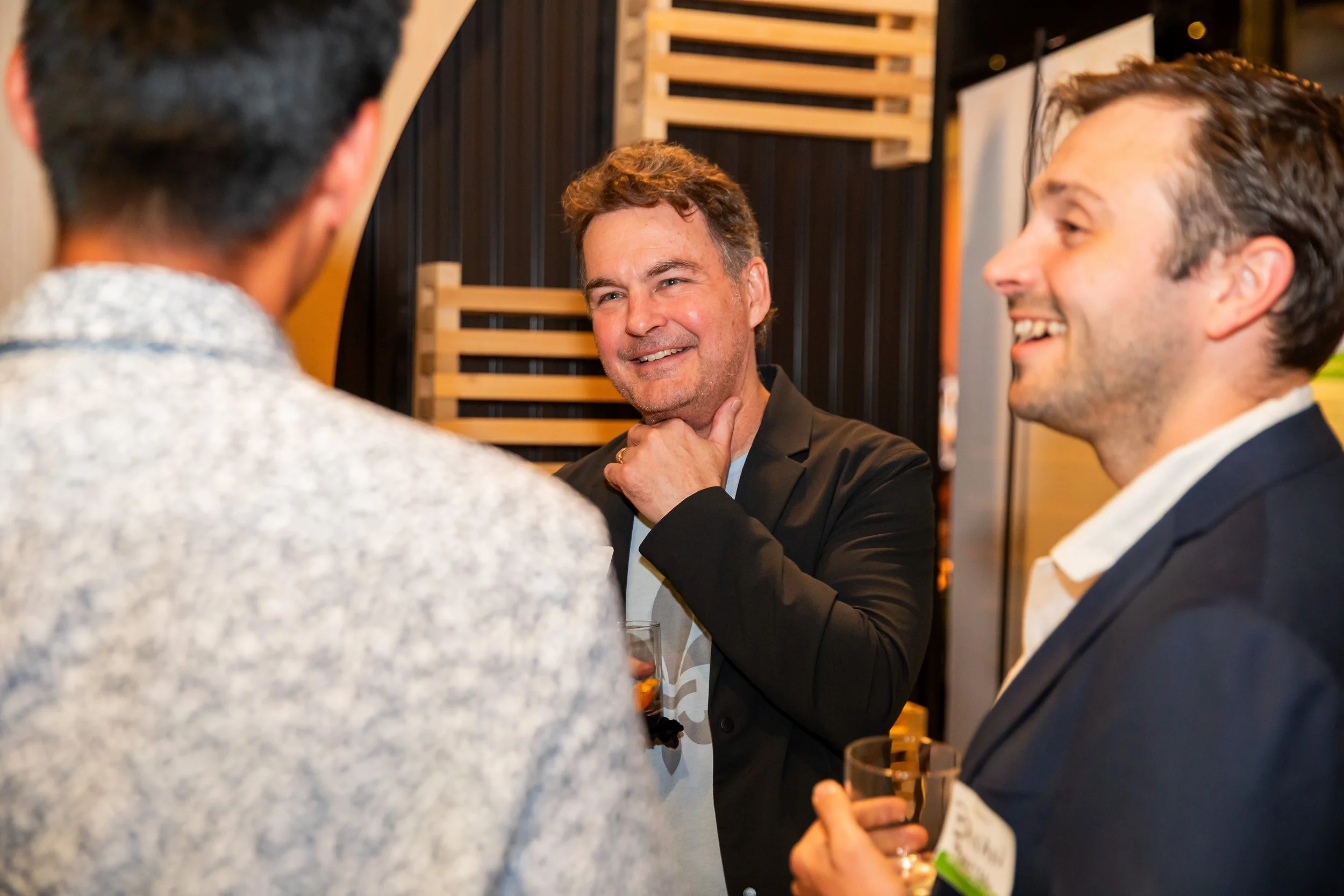 Three men engaged in a conversation at a social event. One man in a black blazer and white shirt is smiling and touching his chin, while another man in a dark suit and white shirt is holding a wine glass and smiling. A third person, partially visible