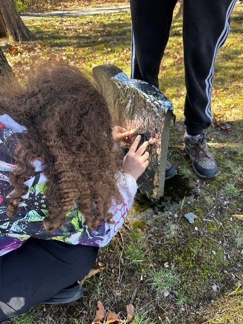 A girl with curly hair crouching in a park, touching a small tombstone, with a person standing nearby.