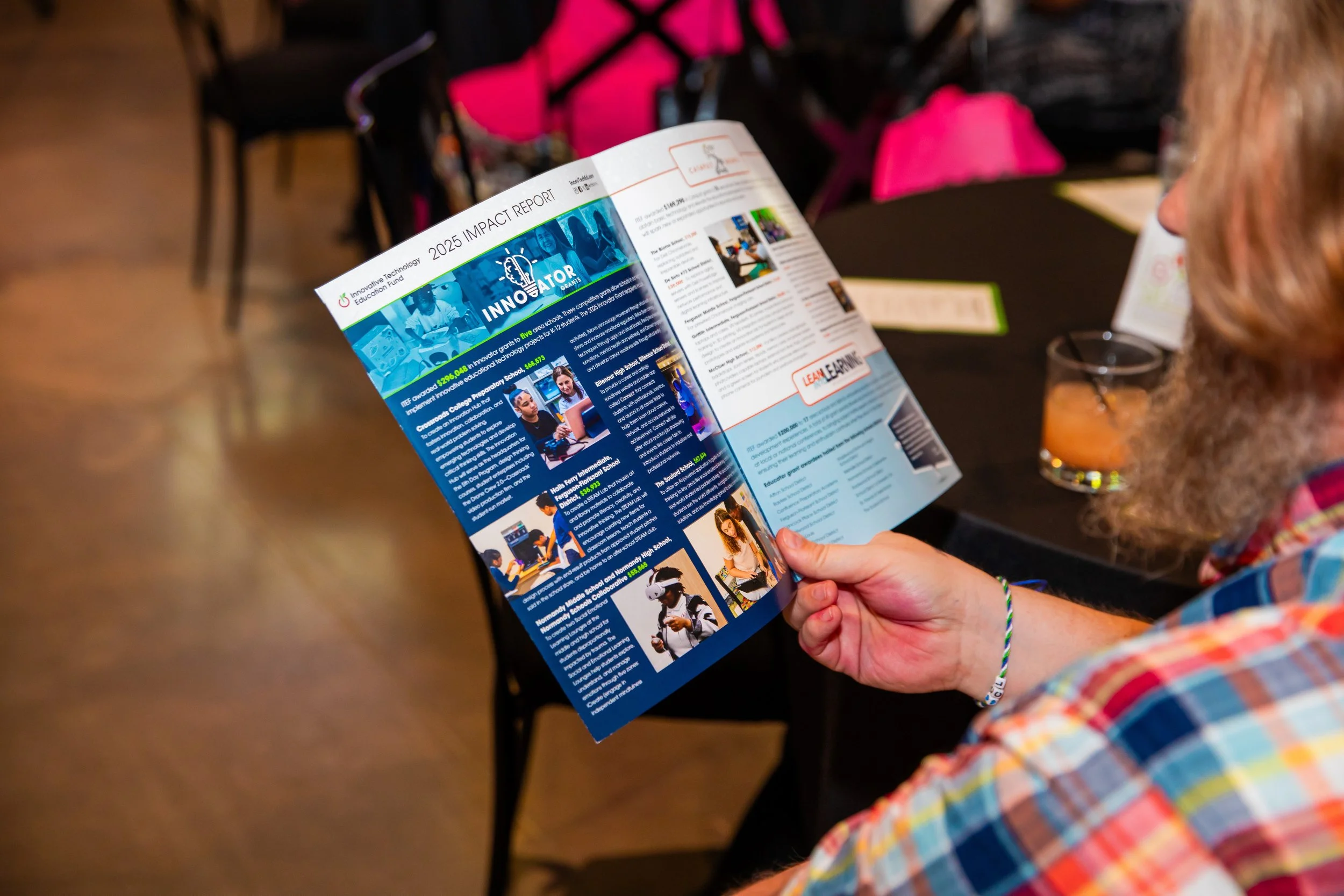 Person holding and reading a colorful printed brochure or magazine with articles and images, seated at a table.