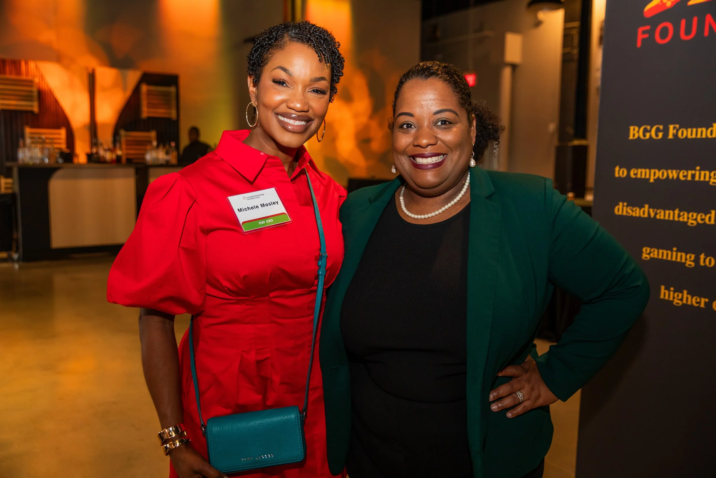 Two smiling women standing close together at an indoor event, one wearing a red jacket with a name tag reading 'Michele Mosley' and the other in a green blazer and pearl necklace, with a black banner in the background.