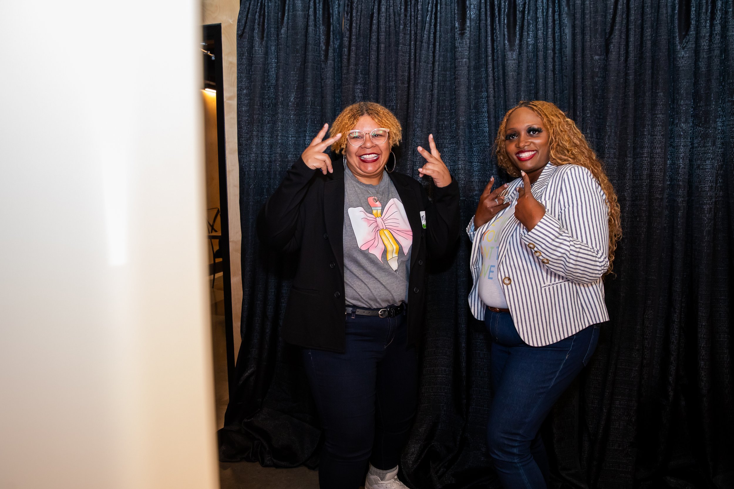 Two women smiling and posing for a photo behind a black curtain at an indoor event. One woman is making a peace sign and wearing glasses, a gray t-shirt with a colorful butterfly graphic, and a black jacket. The other woman is pointing at herself and