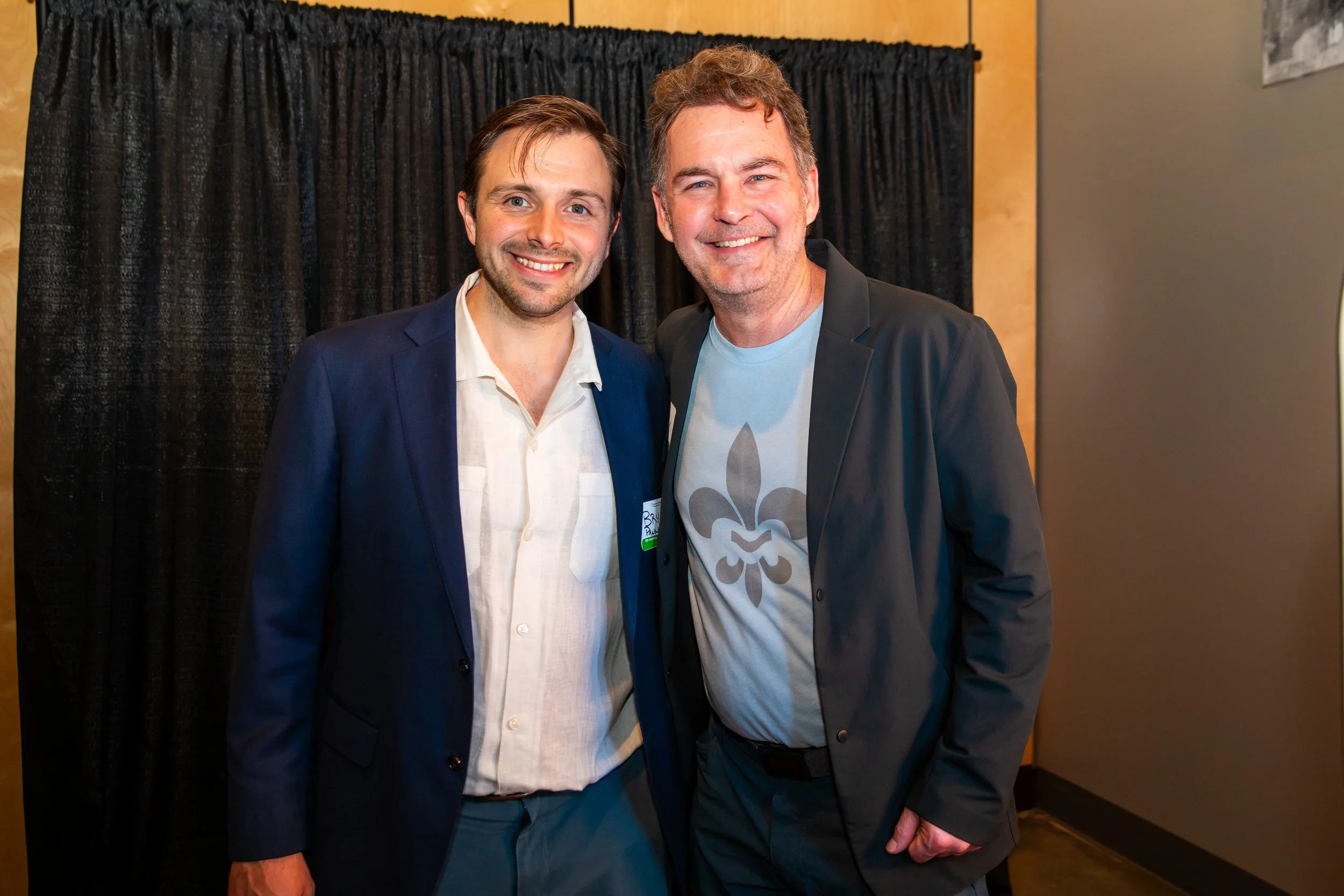 Two men standing close together, smiling at the camera. The man on the left is wearing a navy blazer and a white shirt. The man on the right is wearing a black blazer over a gray T-shirt with a fleur-de-lis design. They are indoors with a black curta