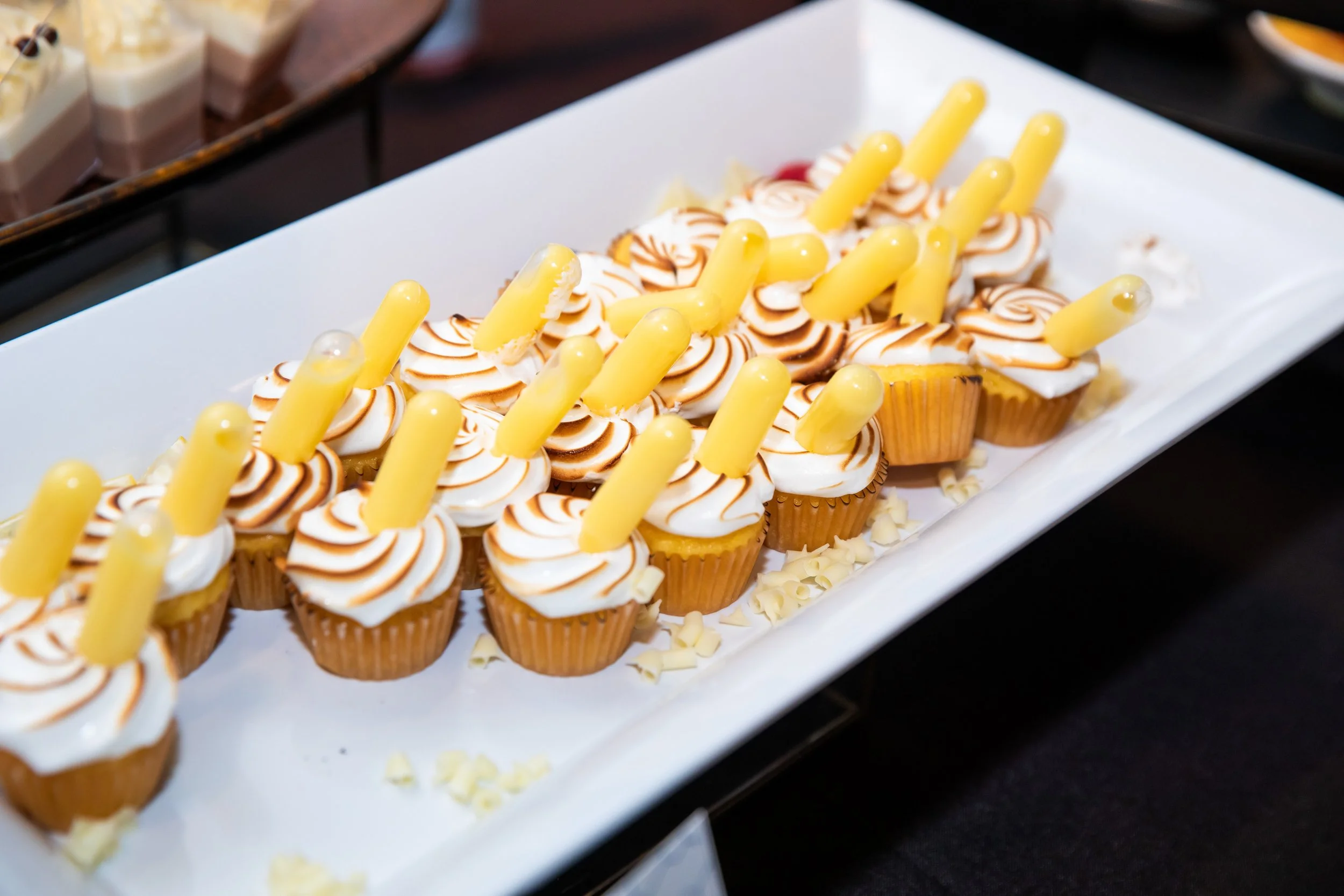 Cupcakes with swirled white and caramel-colored frosting, yellow candle-like decorations on top, on a white tray with white chocolate shavings.