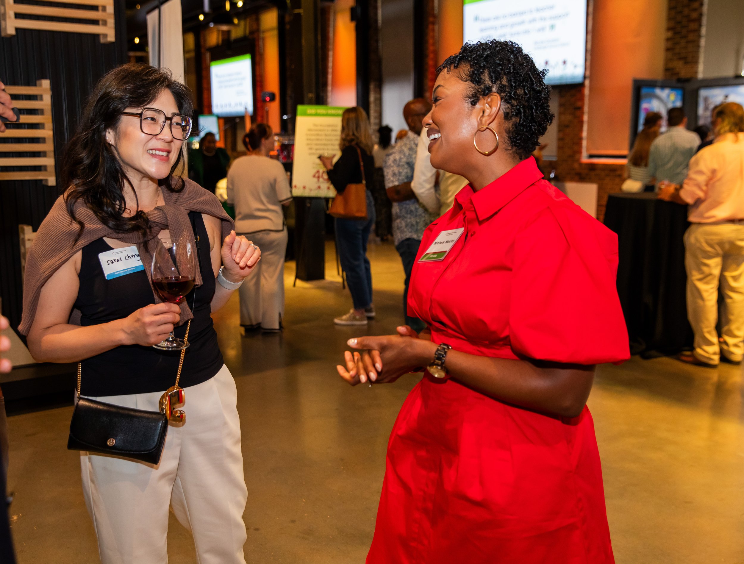 Two women talking at a social event. One with glasses and a name tag, holding a glass of red wine, and the other wearing a red blouse with a name tag, smiling. There are other people in the background and large screens on the walls.