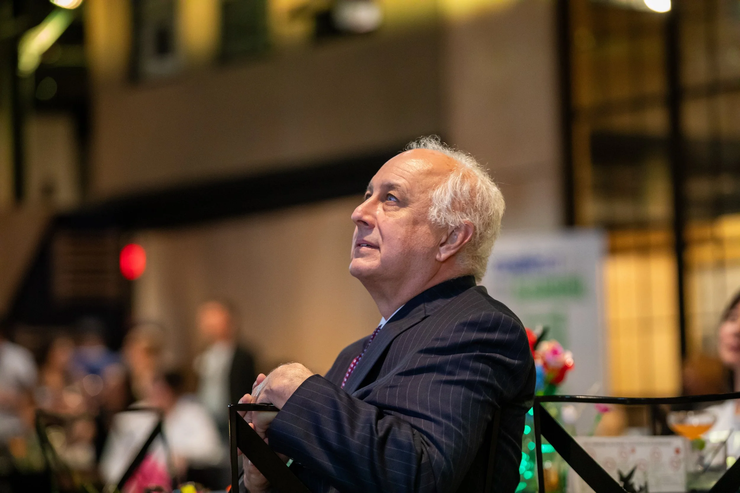 An elderly man with white hair in a pinstripe suit looks upward while sitting at a formal event with blurred people and colorful decorations in the background.