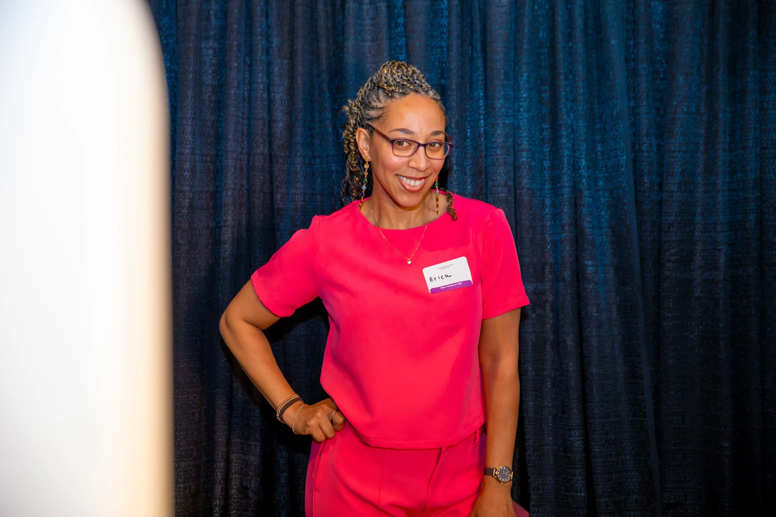 A woman with glasses, gold earrings, and a name tag that reads 'Erica' is standing in front of a dark blue curtain, smiling, and wearing a bright pink outfit with her hand on her hip.
