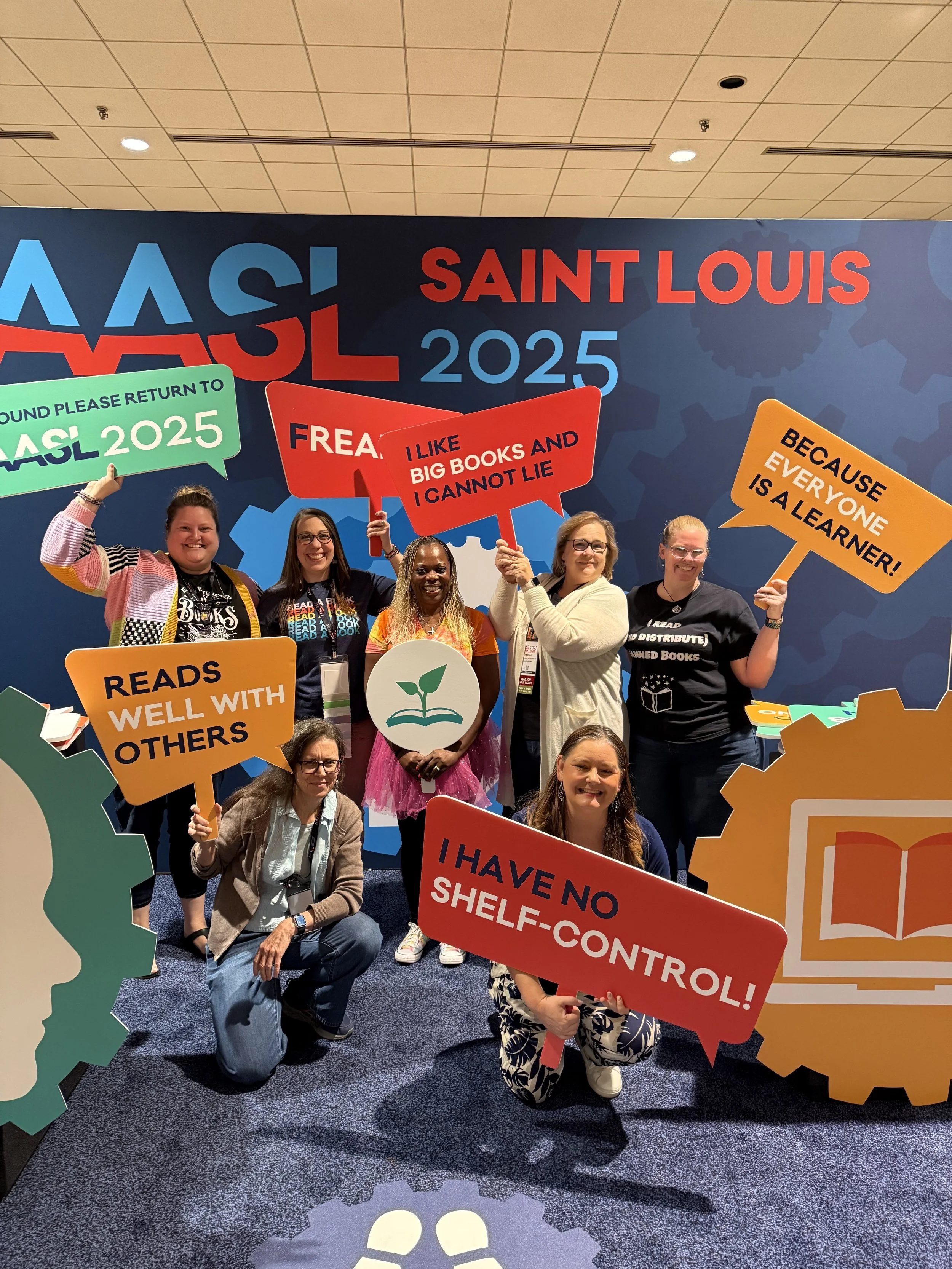 Group of diverse women at a conference holding colorful speech bubble signs with inspirational messages, standing in front of a large wall display that reads "VASL 2025" and "Saint Louis 2025" with gear designs in the background.