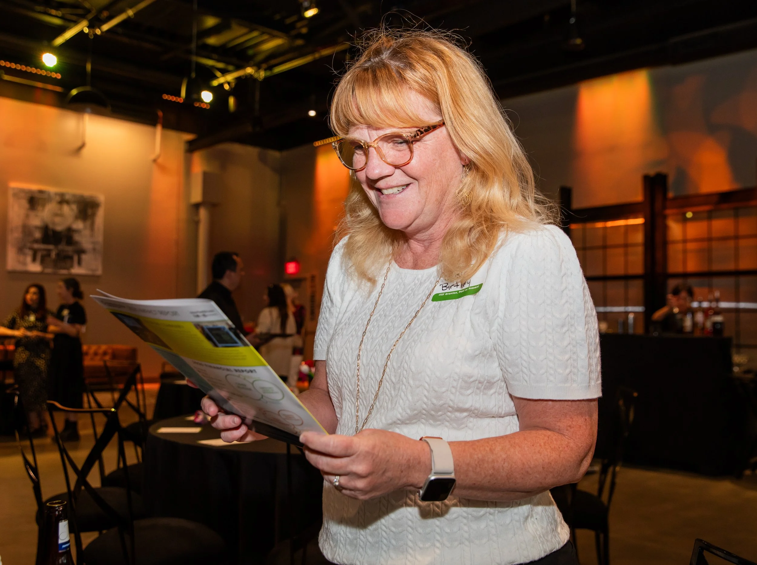 A woman with red hair, glasses, and a white textured shirt is smiling and reading a pamphlet at an indoor event. There are people and tables in the background with warm lighting.
