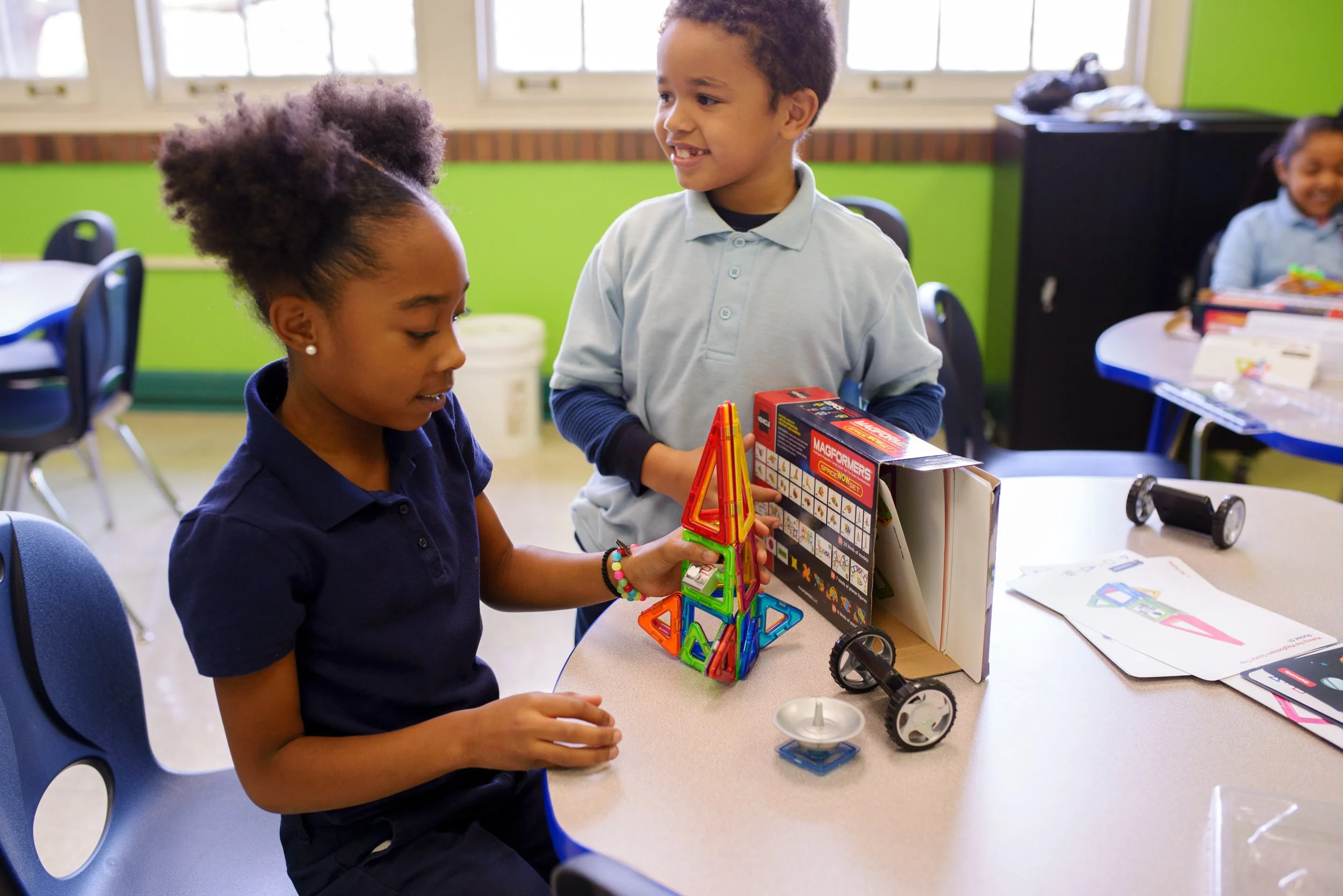 Two children, a girl and a boy, are working together at a table in a classroom. The girl is seated and looking at the tower they built with colorful magnetic pieces. The boy stands next to her, smiling and holding a board game box. Other children are visible in the background, and the classroom has green walls and large windows.