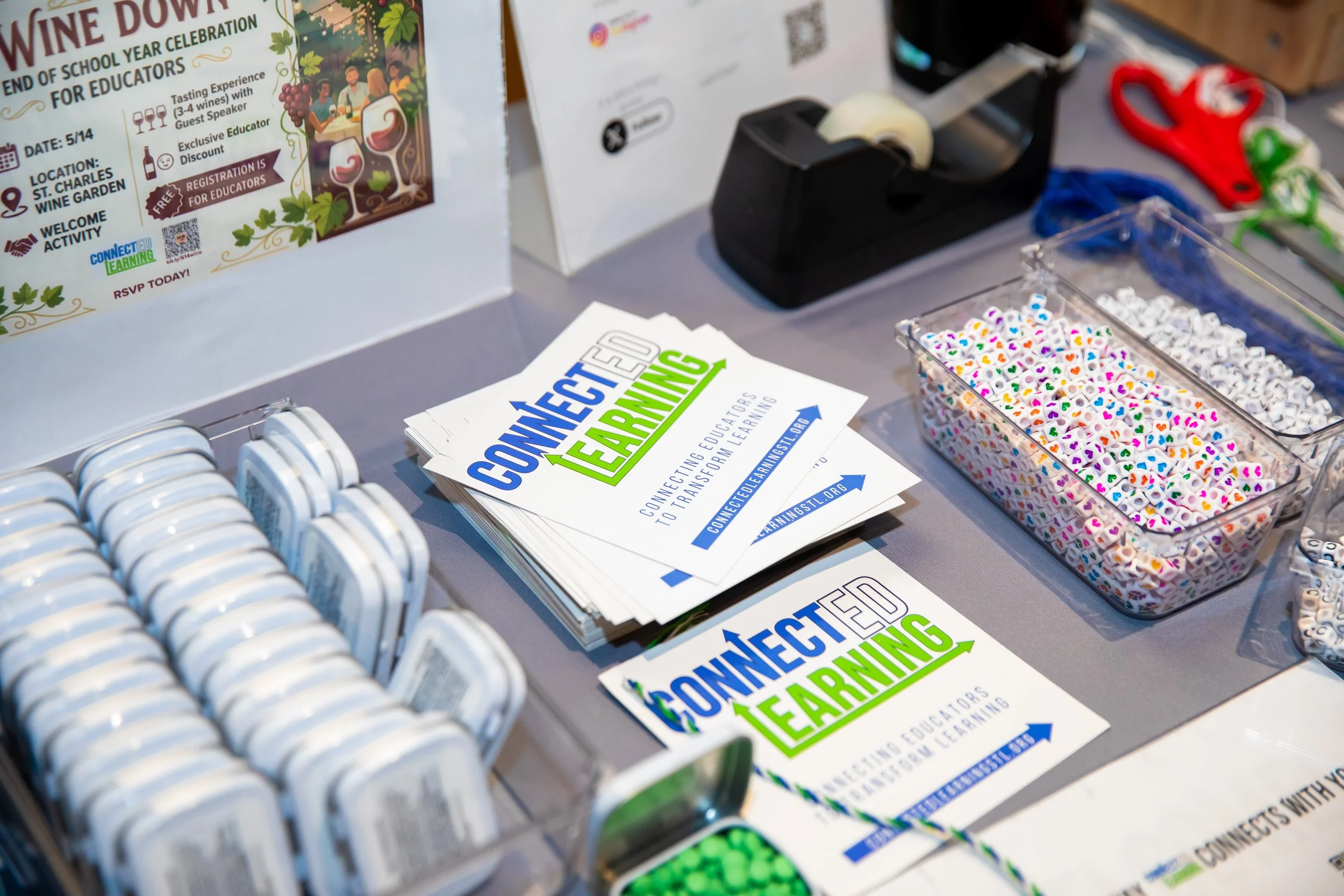A table with promotional materials for connecting educators through learning, featuring brochures, a container of colorful heart-patterned beads, a tape dispenser, and a sign about a wine tasting event for educators.