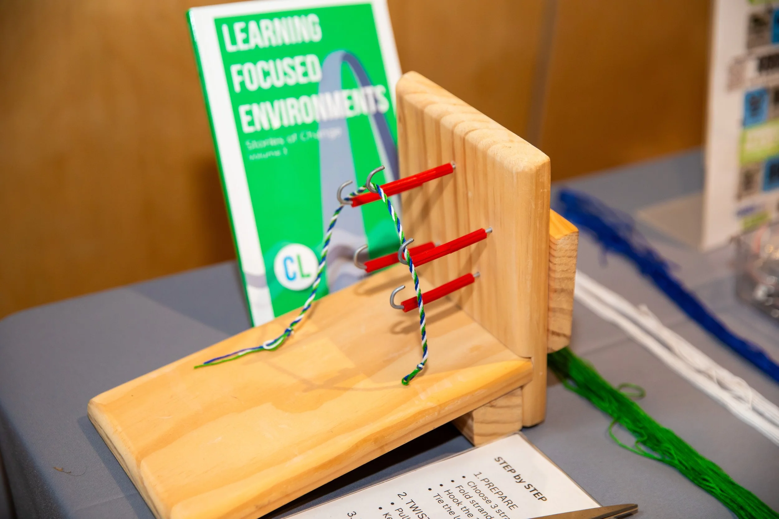 A wooden display stand with colored hooks holding a twisted green, blue, and white string, next to a green instructional sign about environmental learning, with a blurred background of a room.