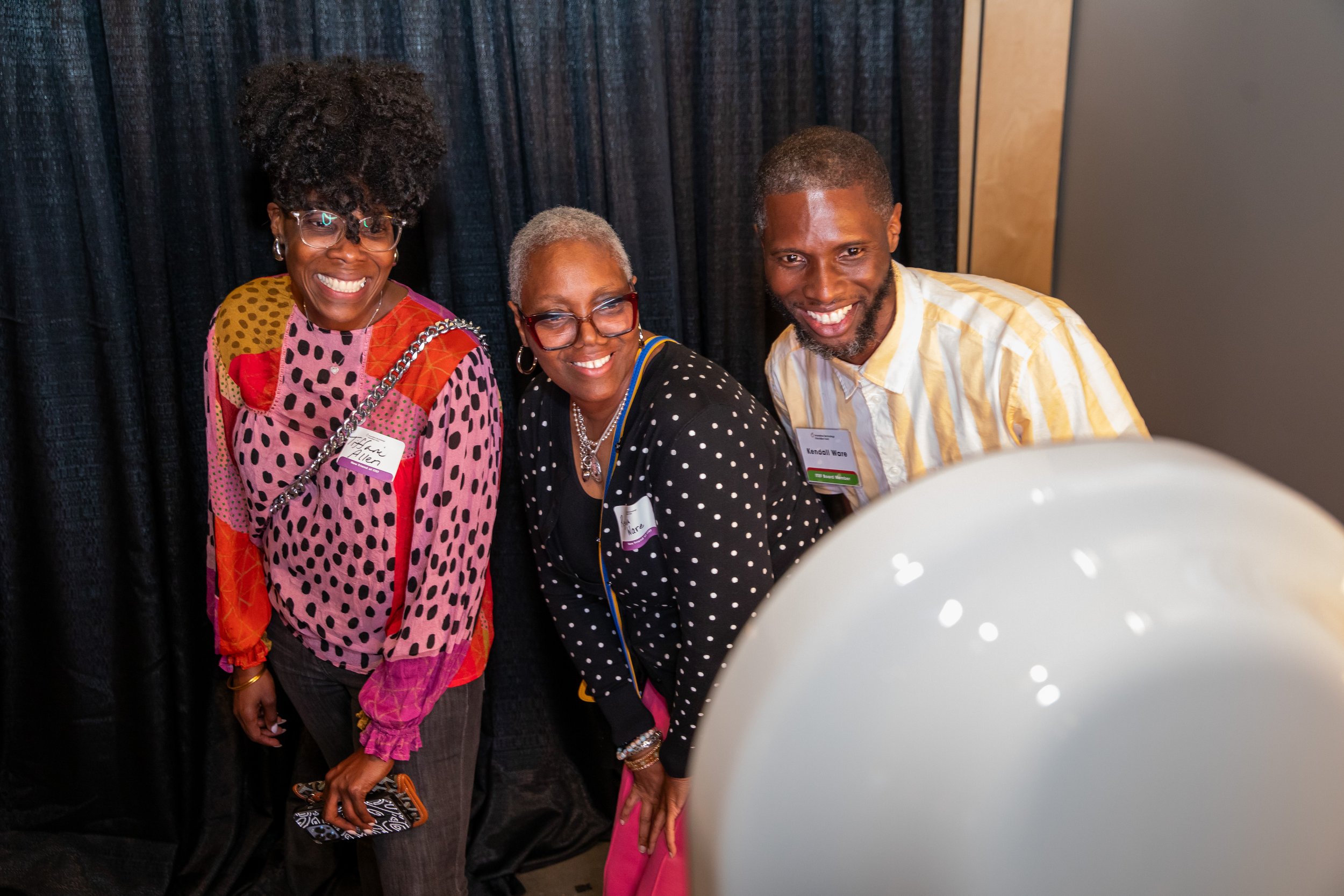 Three people smiling and posing for a photo in front of a black curtain, with a white balloon near the camera.