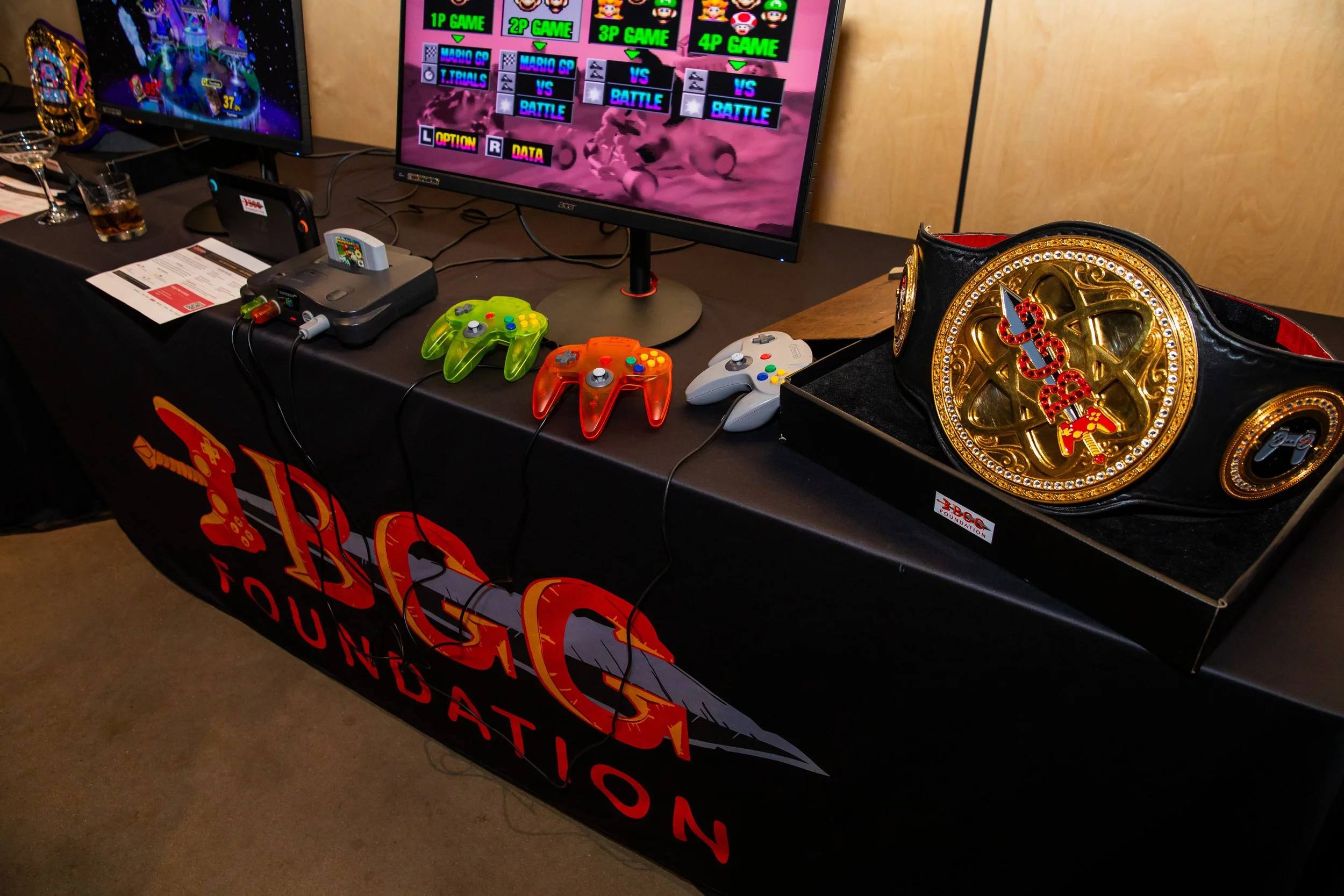 A gaming setup display features a large monitor showing a colorful video game menu, with two other monitors visible in the background. On the table, there are multiple game controllers including a green, orange, and white one, a Game Boy Advance SP, 