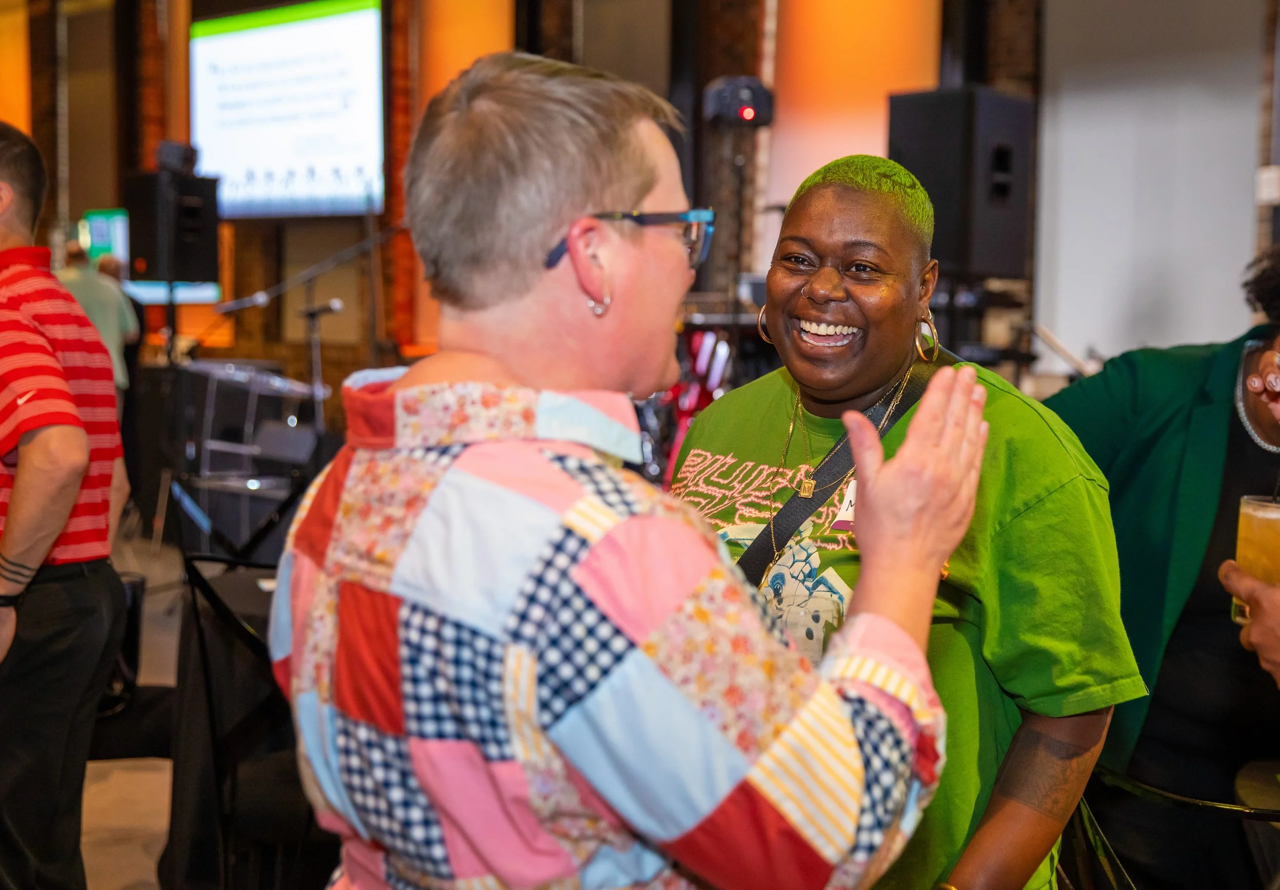 Two women are engaged in a cheerful conversation at a social gathering. One woman has short, light-colored hair and glasses, while the other has a vibrant green hairstyle and is smiling broadly. They are standing close to each other, with a lively ba