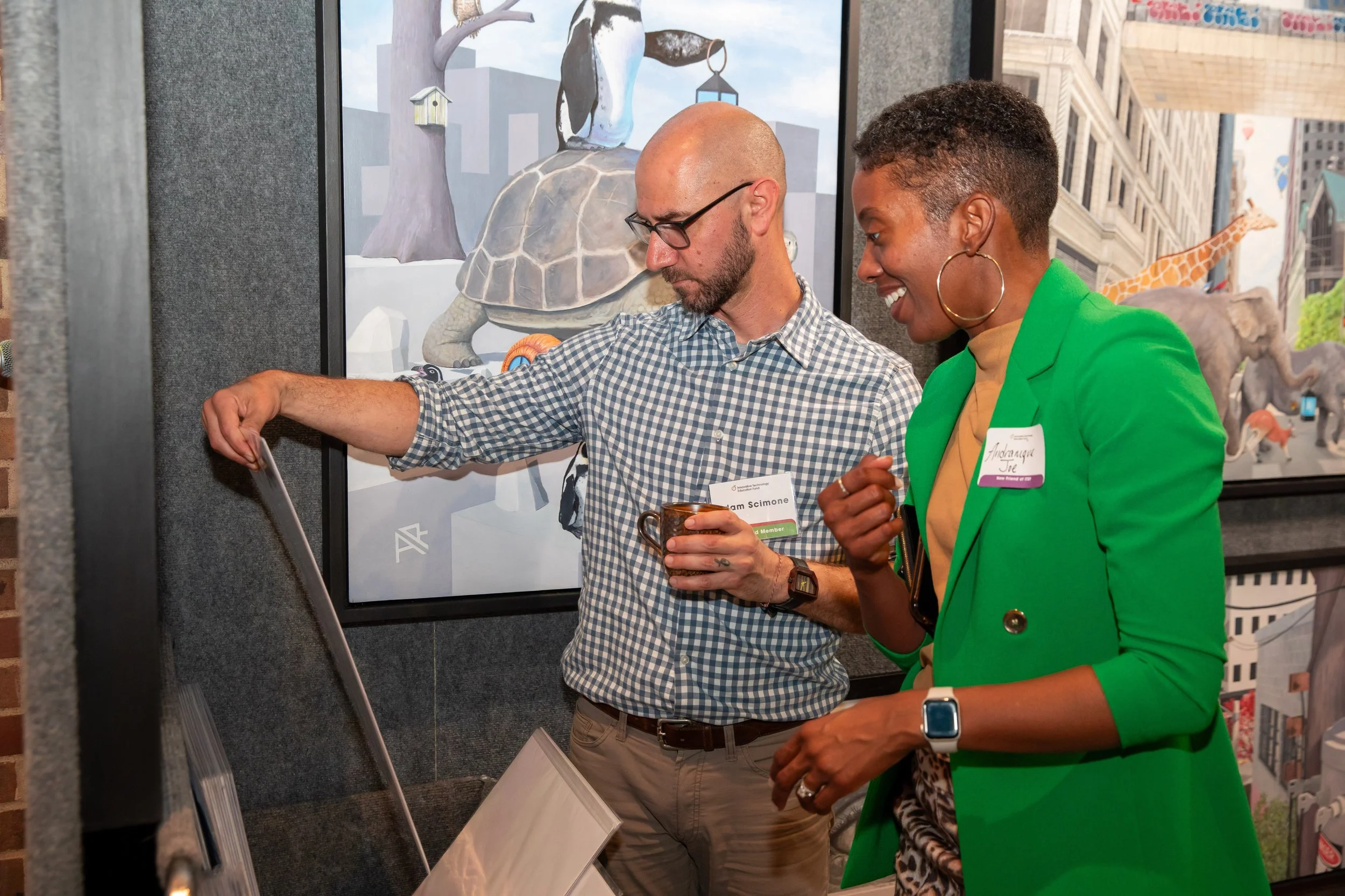 Two people, a man and a woman, are engaging in conversation or an activity at an art exhibit. The man is wearing a checkered shirt, glasses, and a watch, holding a mug, and looking at a display. The woman is smiling, wearing a bright green blazer, la