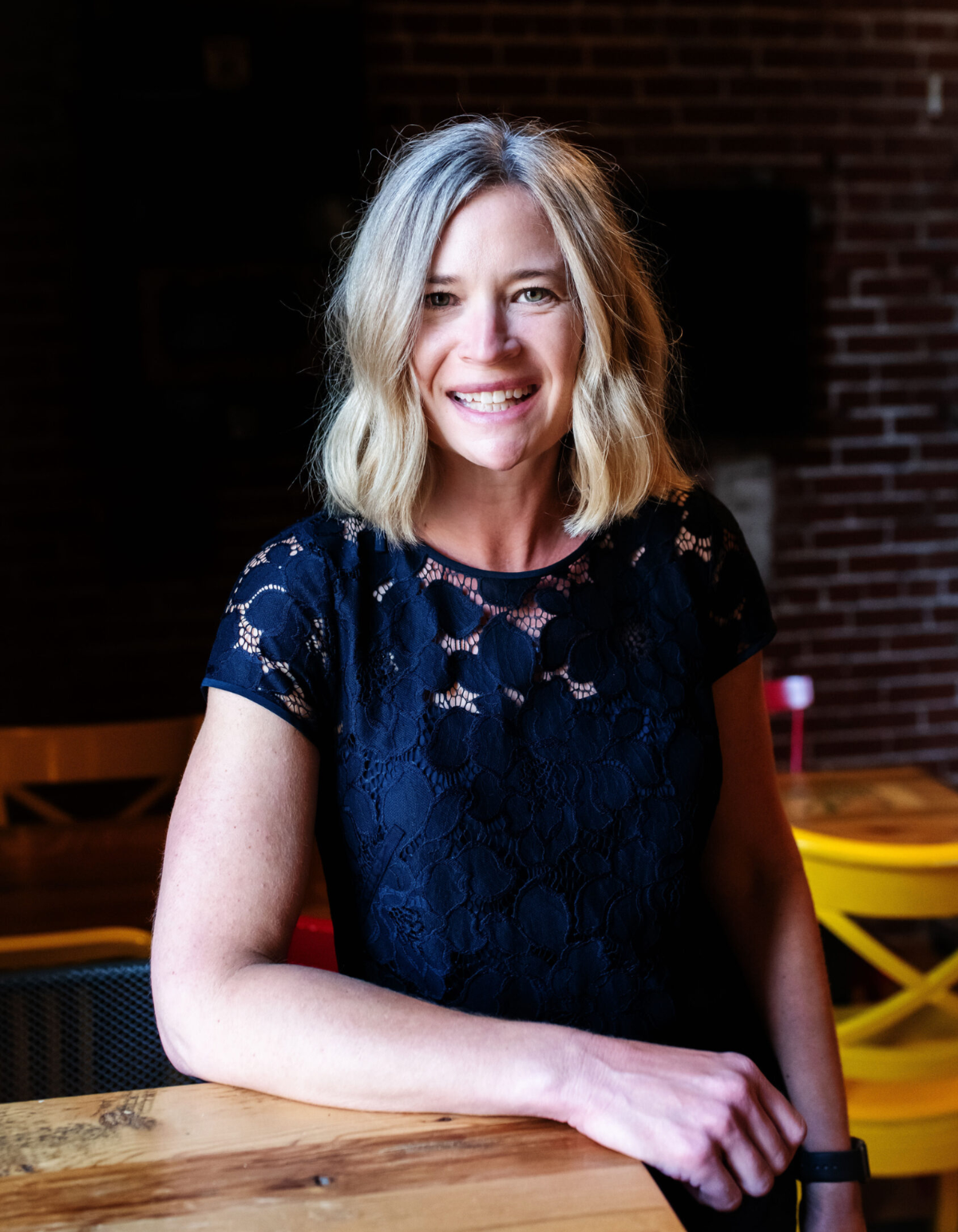 A smiling woman with blonde hair, wearing a black lace top, sitting at a wooden table in a dimly lit room with brick walls.