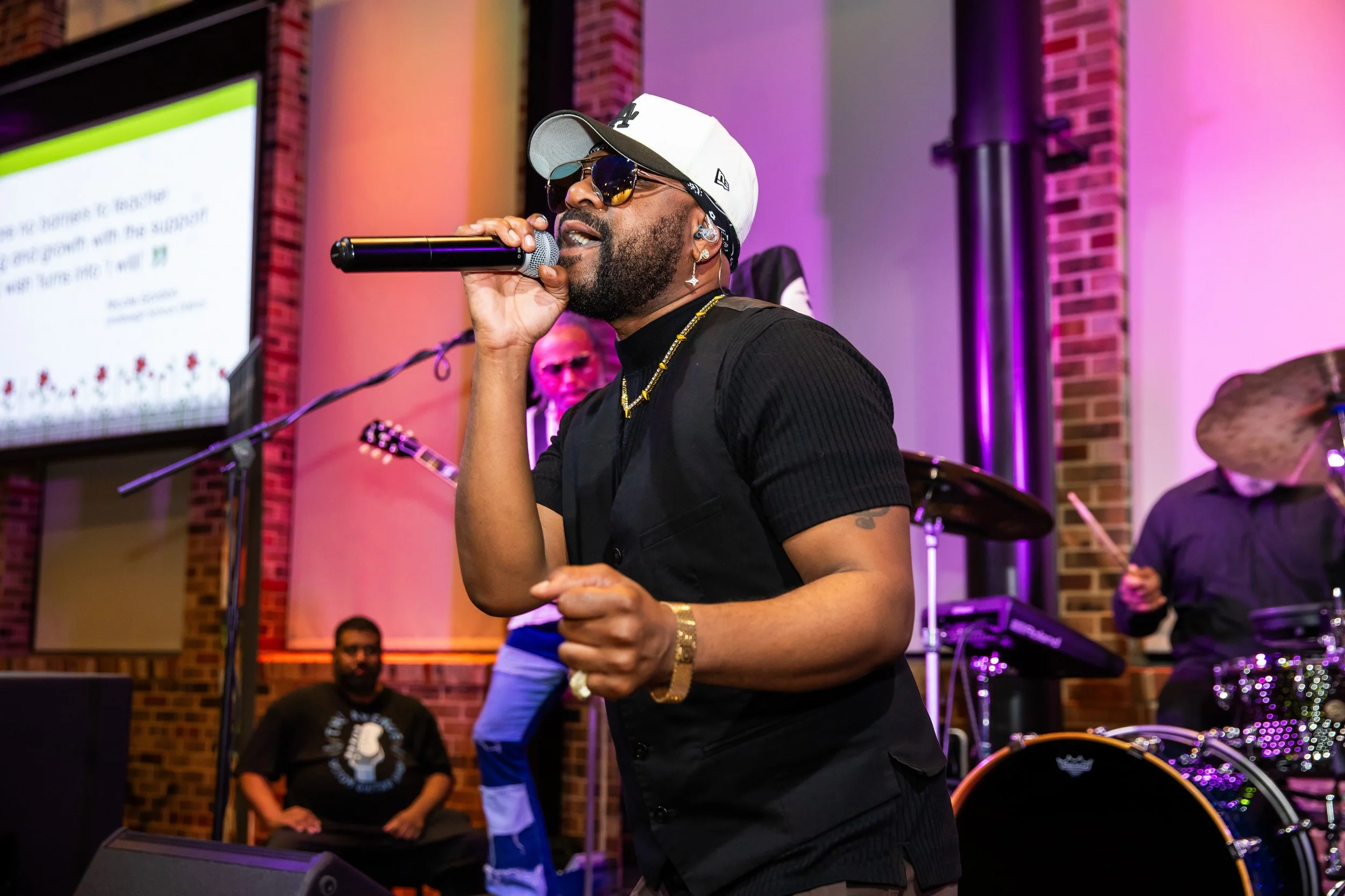 A male singer wearing sunglasses, a white cap, earrings, and a black shirt performs with a microphone on stage in front of a colorful background with purple and pink lighting.