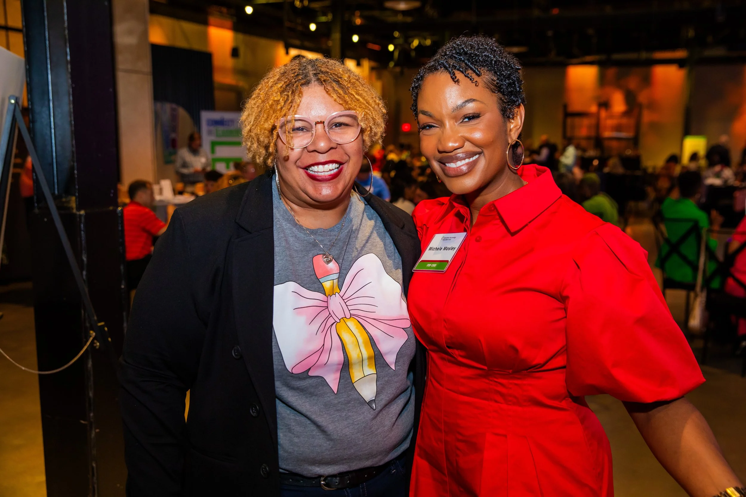 Two women smiling at an indoor event, one wearing glasses and a black blazer with a gray T-shirt featuring a pink loveheart design, the other in a red blouse with a name tag.