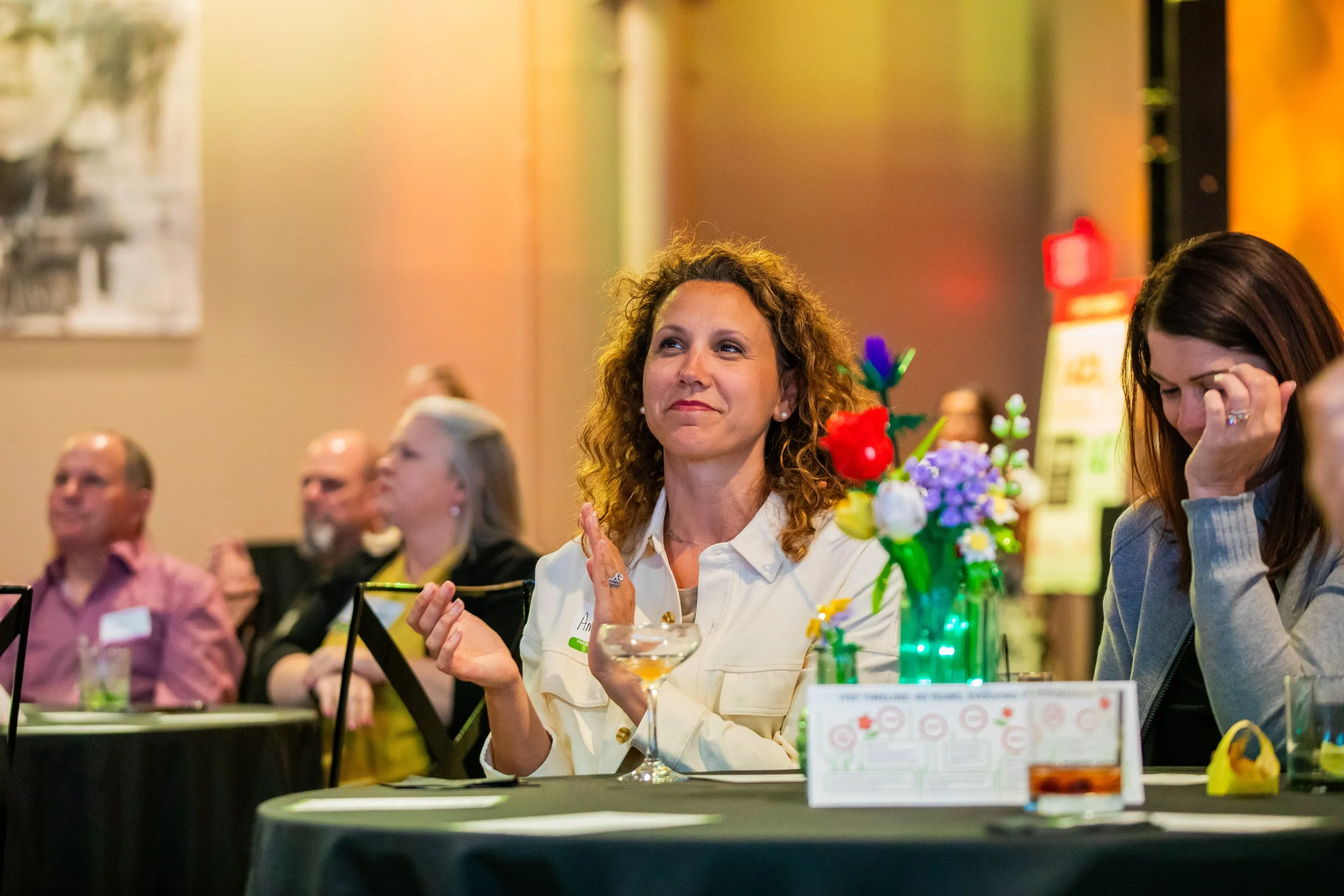 A group of people seated at a table during a social event, with a woman with curly hair smiling and clapping in the center, colorful decorations and a framed picture on the wall behind them.