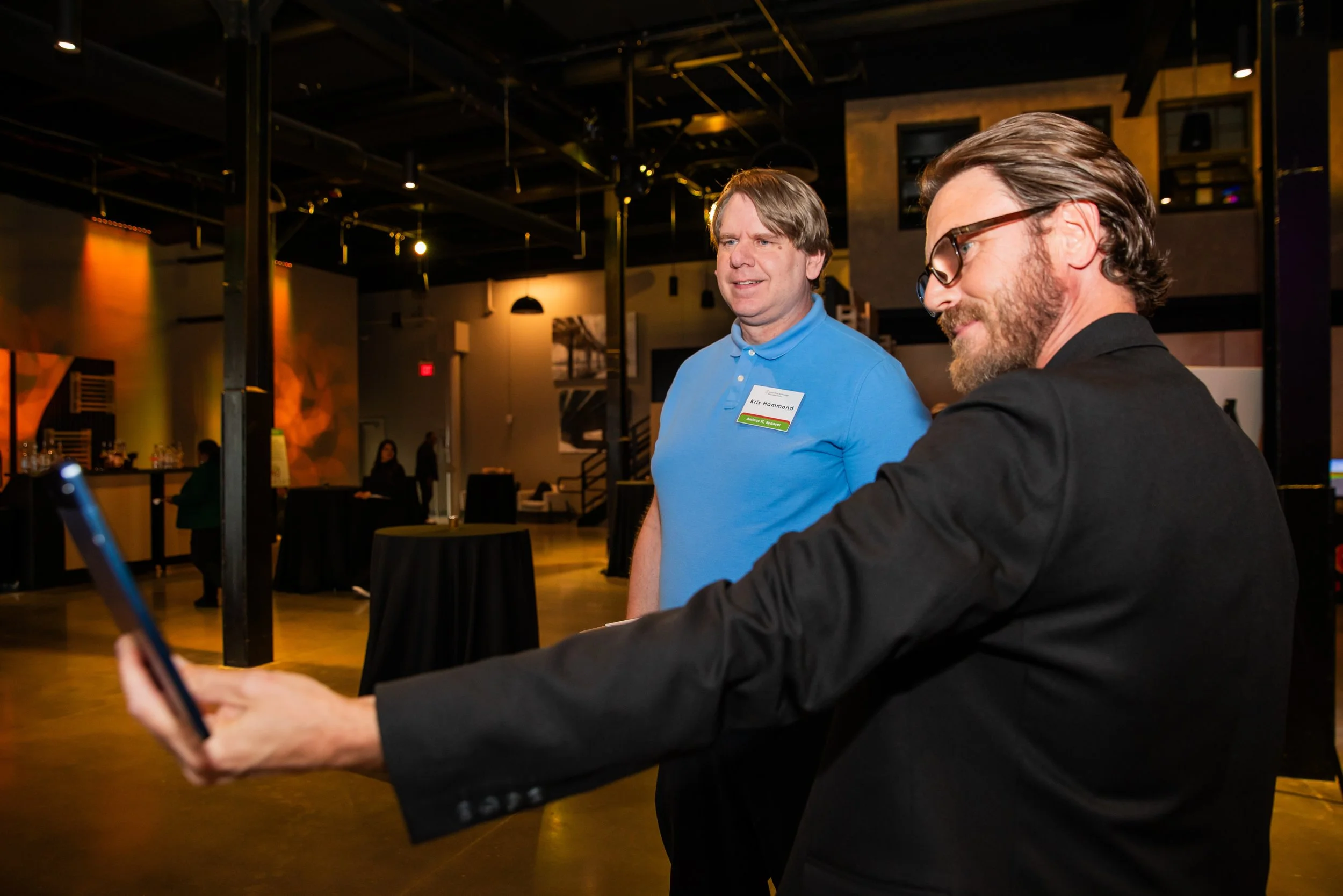 Two men are standing at an indoor event or gathering, with one man smiling and wearing a blue polo shirt with a name tag that reads "Kris Hammond," and the other man looking at his phone, wearing glasses and a black suit. The background shows a moder