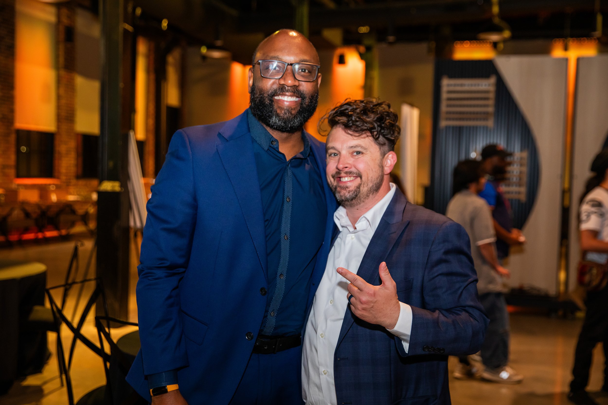 Two men in formal attire smiling at an event, with one pointing at the other, in a warmly lit indoor space with other people in the background.