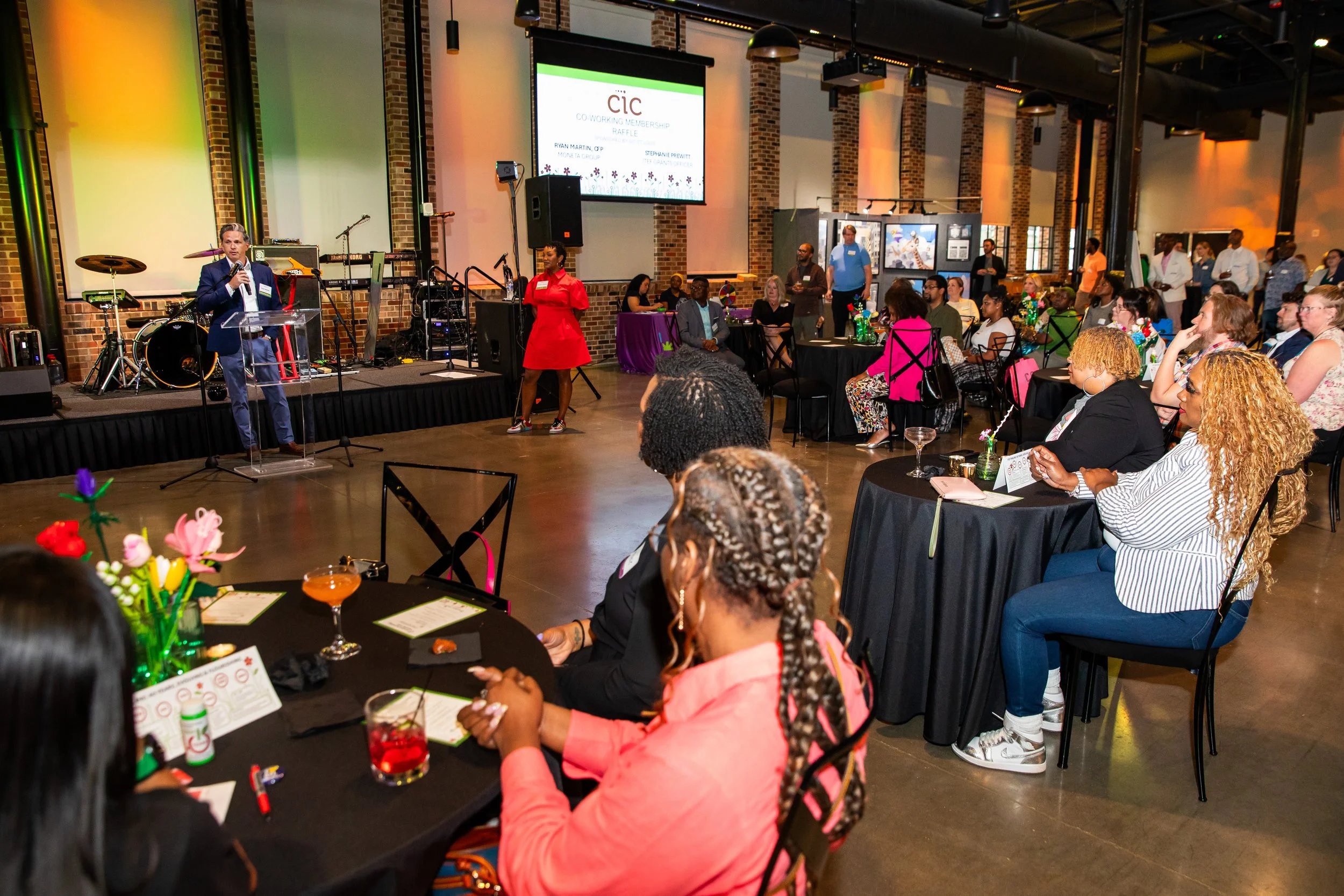 A speaker at a podium with a microphone addressing an audience at an event called CIC Co-working Membership Raffle. The event is held in a spacious industrial-style venue with brick walls, large windows, and colorful lighting. The audience is seated 